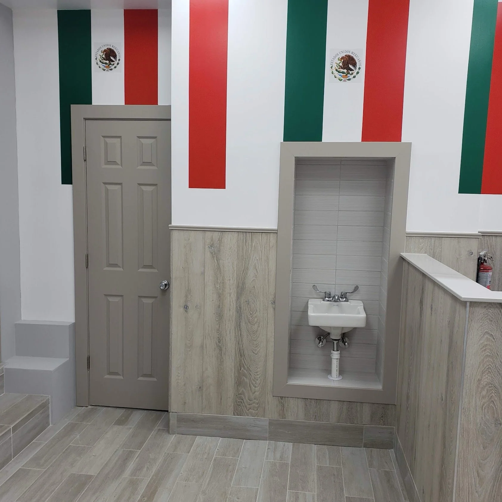 A wall with Mexican flags painted on it, two doors, a small sink, and a fire extinguisher inside a room.