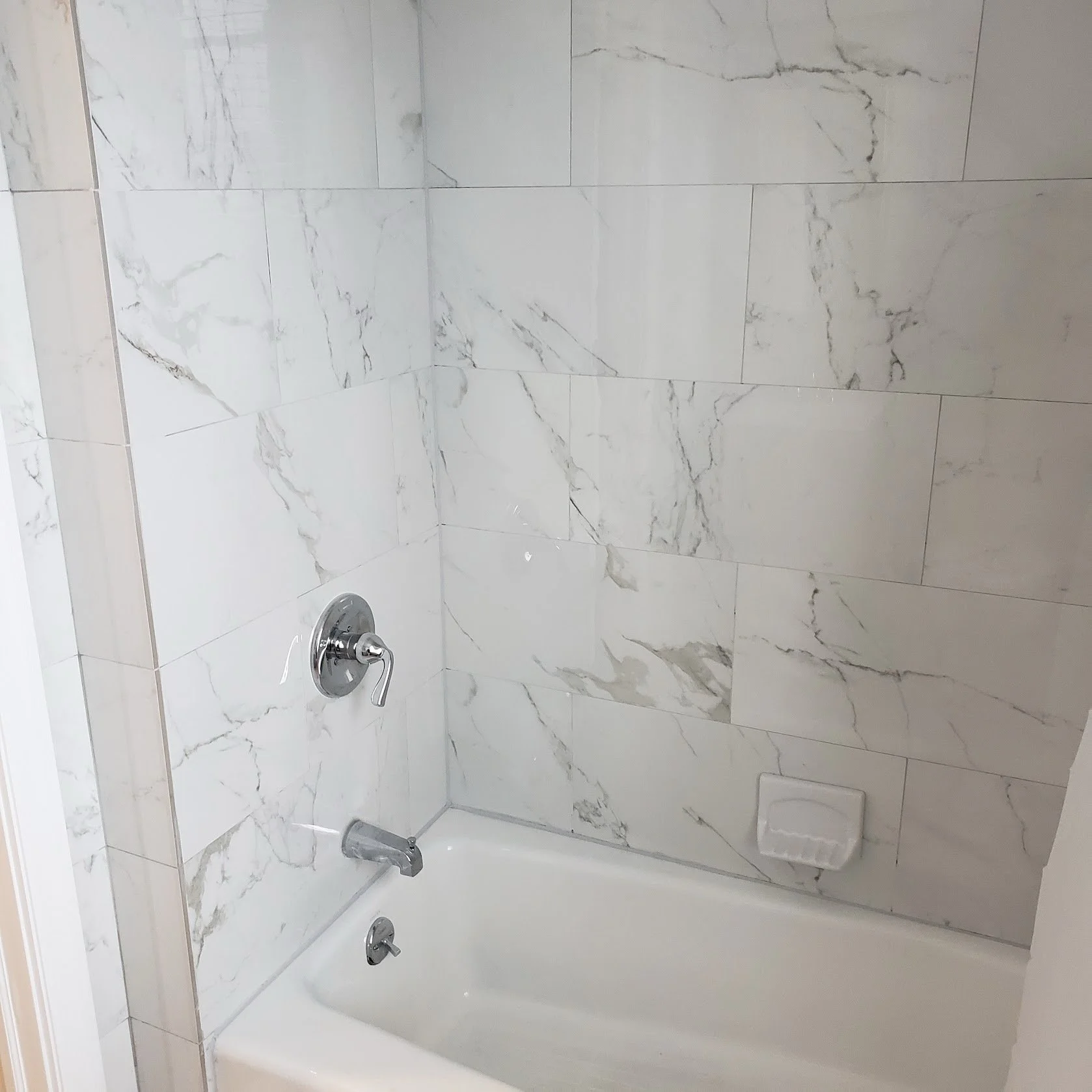 A marble tiled bathtub/shower area with a silver handle, a faucet, and a showerhead, with a vent cover in the corner.