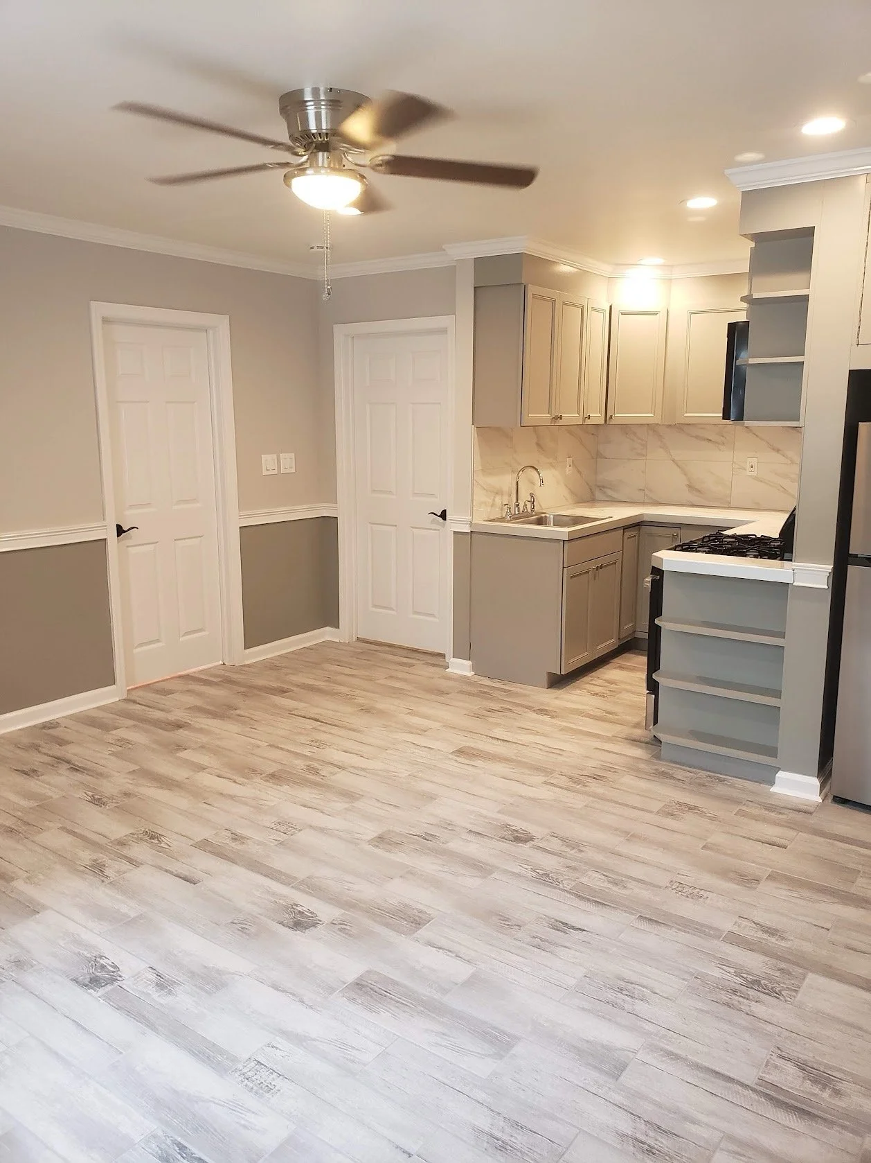 Empty living room and kitchen with light wood flooring, gray cabinets, white walls, and a ceiling fan.