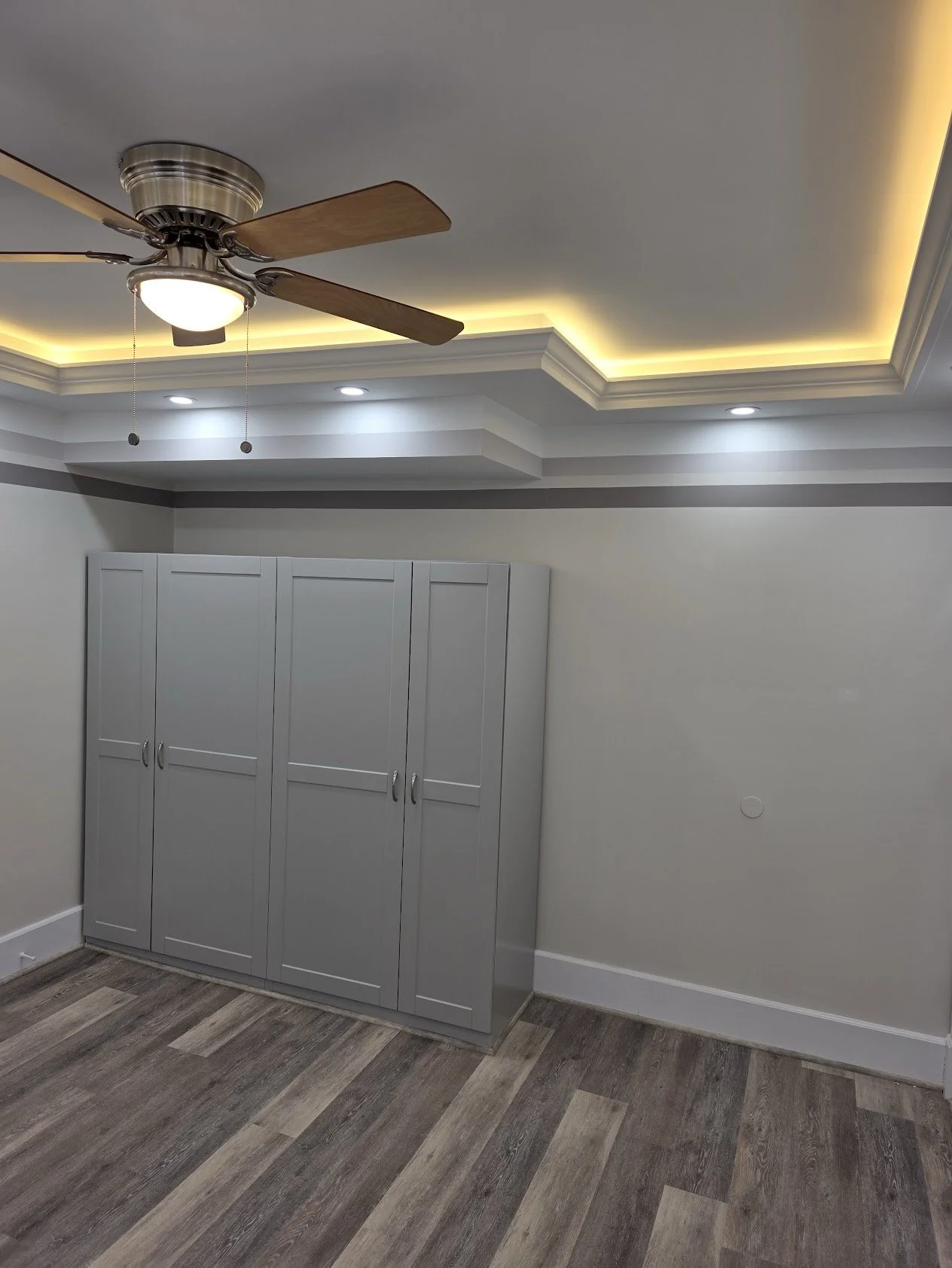 A room with a ceiling fan, recessed lighting, and a white closet door. The ceiling has a tray design with LED strip lighting around the edges. The floor has wood-style laminate flooring.