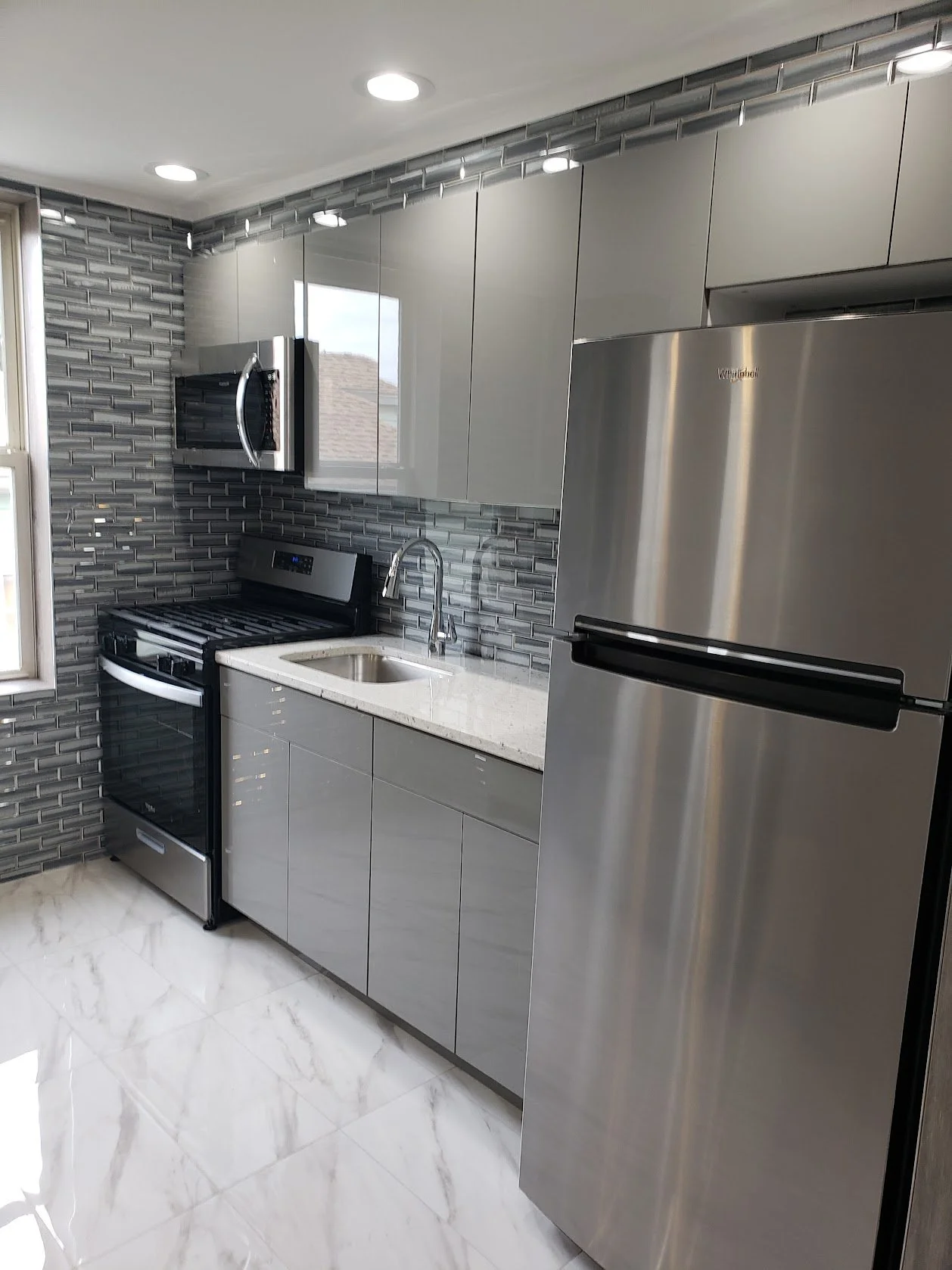Modern kitchen with gray cabinets, stainless steel refrigerator, microwave, stove, and gray tiled backsplash. Marble floor and a window next to the stove.