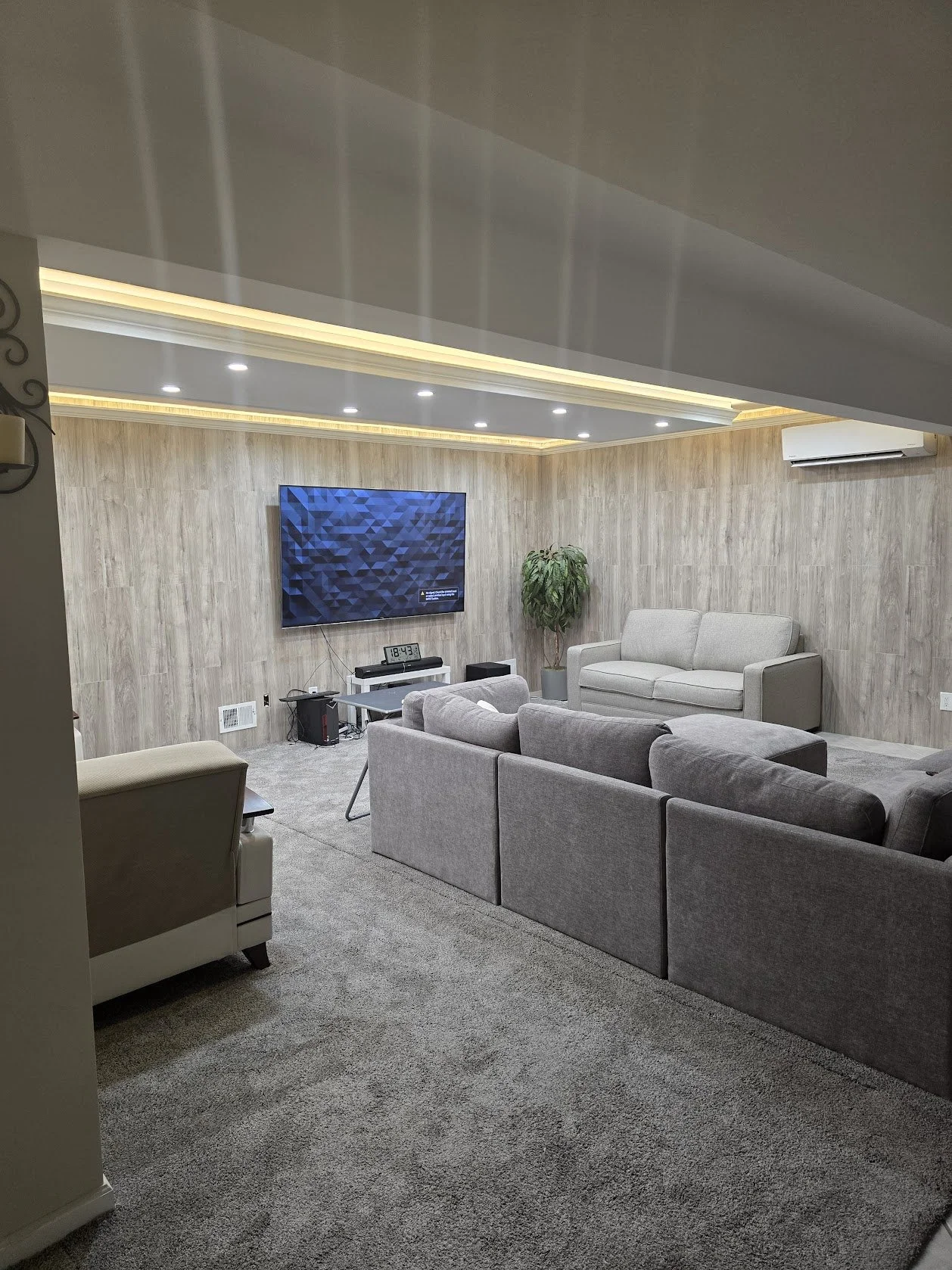 A modern living room with a gray sectional sofa, a beige armchair, a small white table, and a large flat-screen TV mounted on a wooden-paneled wall. There's a potted green plant near the TV and ceiling lights with some recessed lighting. Carpeted flo