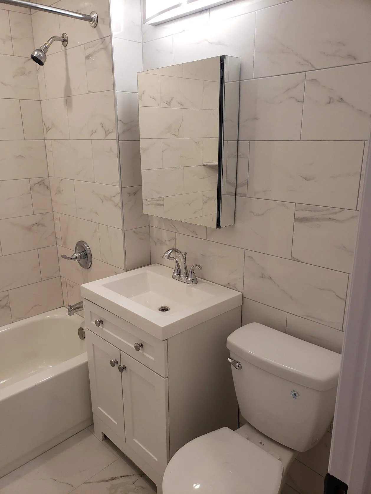 A small bathroom with beige marbled tiles, a shower with a shower head, a small white vanity with a sink and cabinet, a mirrored medicine cabinet, and a toilet.