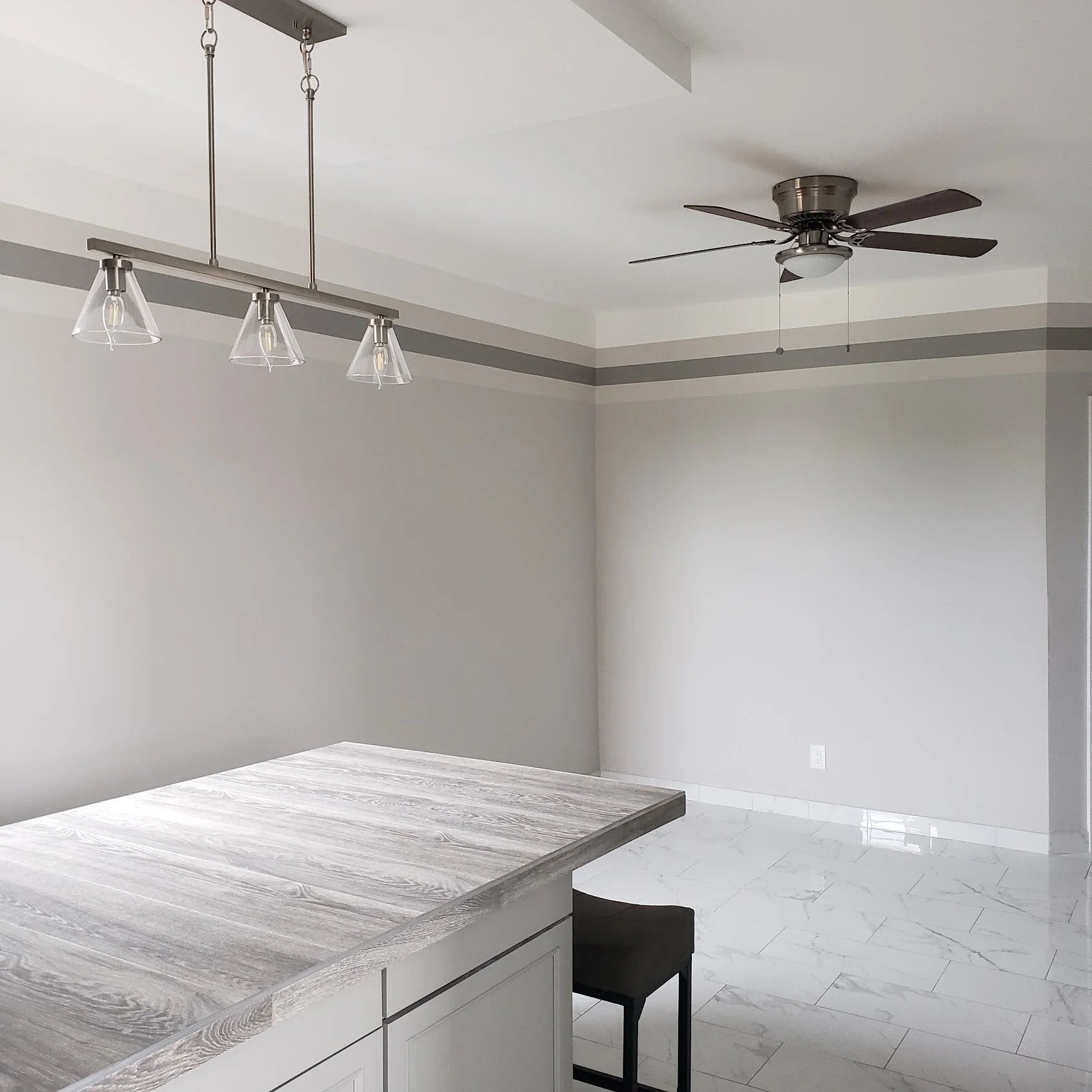 A modern kitchen with white cabinets, a wooden countertop, a silver ceiling fan, a grey and white wall with a decorative stripe, and marble tile flooring.