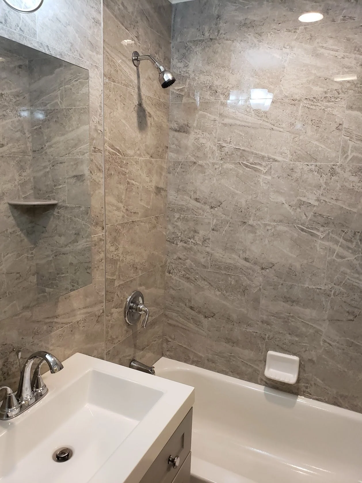 Bathroom with a shower area that has a beige marble tiled wall, a bathtub with a white ceramic sink and chrome faucet in the foreground, a soap dish on the bathtub wall, and a shower head.