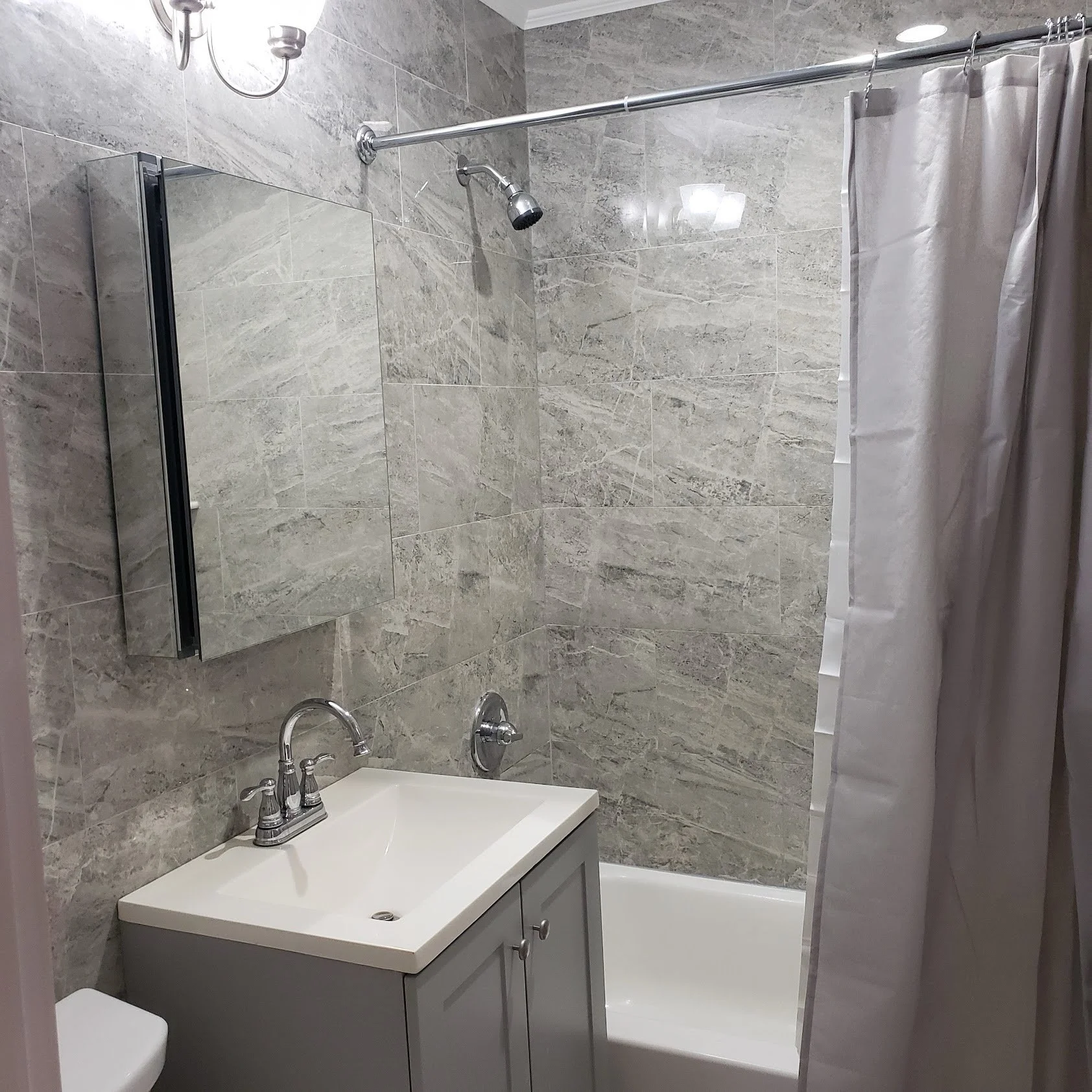 Small bathroom with gray tiled walls, a white sink with a gray cabinet below, a toilet, a bathtub with a shower curtain, a wall-mounted mirror, a light above the mirror, and a showerhead.