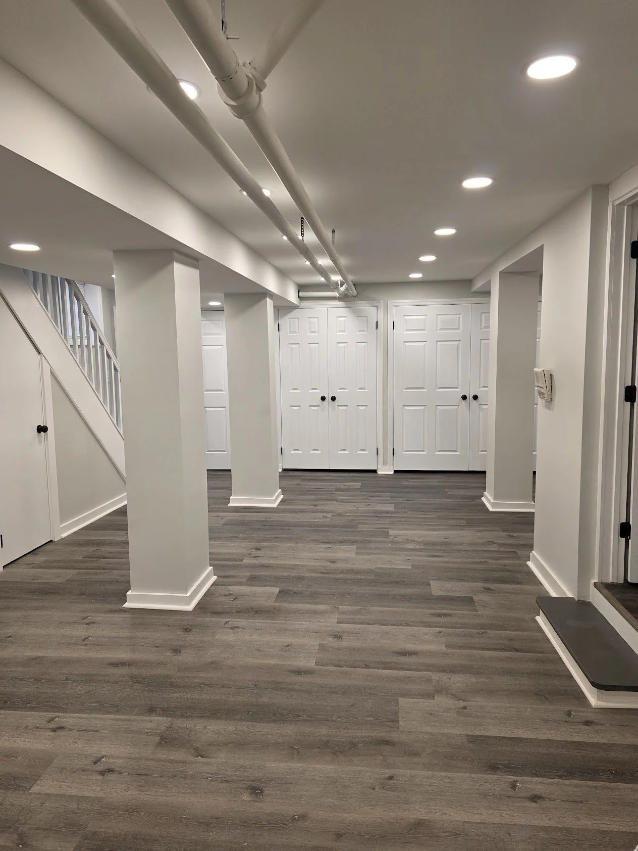 Interior view of a basement with white walls, white doors, and gray wooden flooring, featuring support columns and built-in storage cabinets.