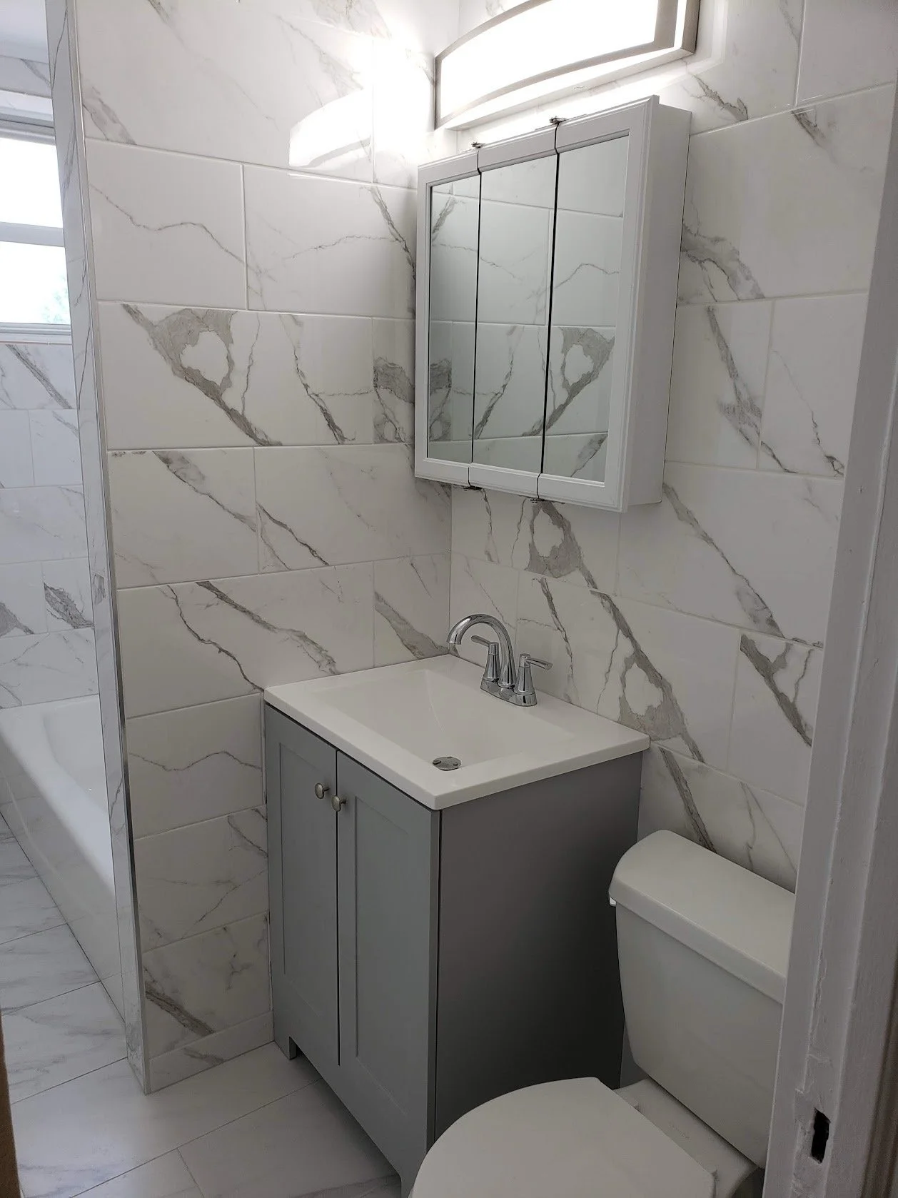 Bathroom with marble tile walls, a small gray vanity with a white sink, a mirror cabinet, and a toilet.