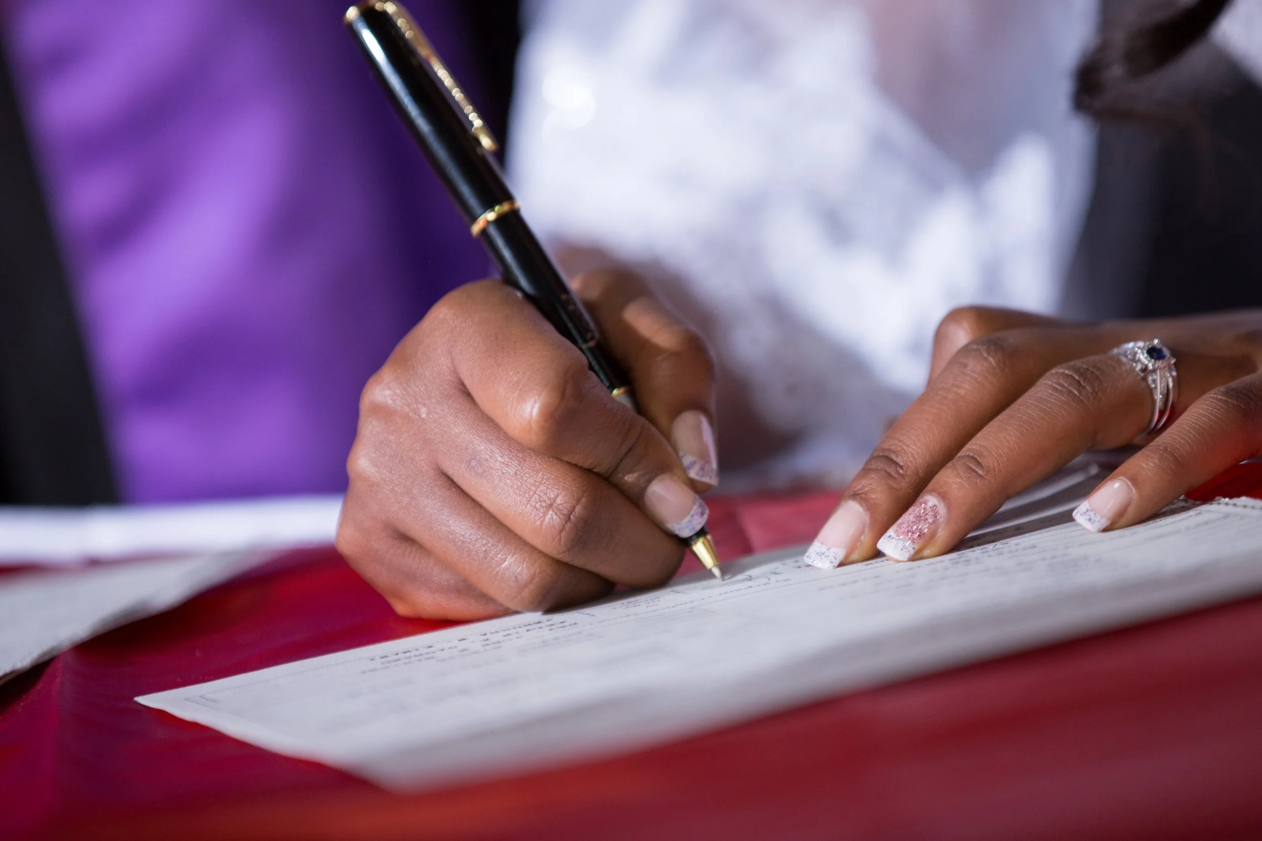 Close-up of a person's hand holding a gold and black pen, writing on a piece of paper, with a ring on one finger and well-manicured nails.