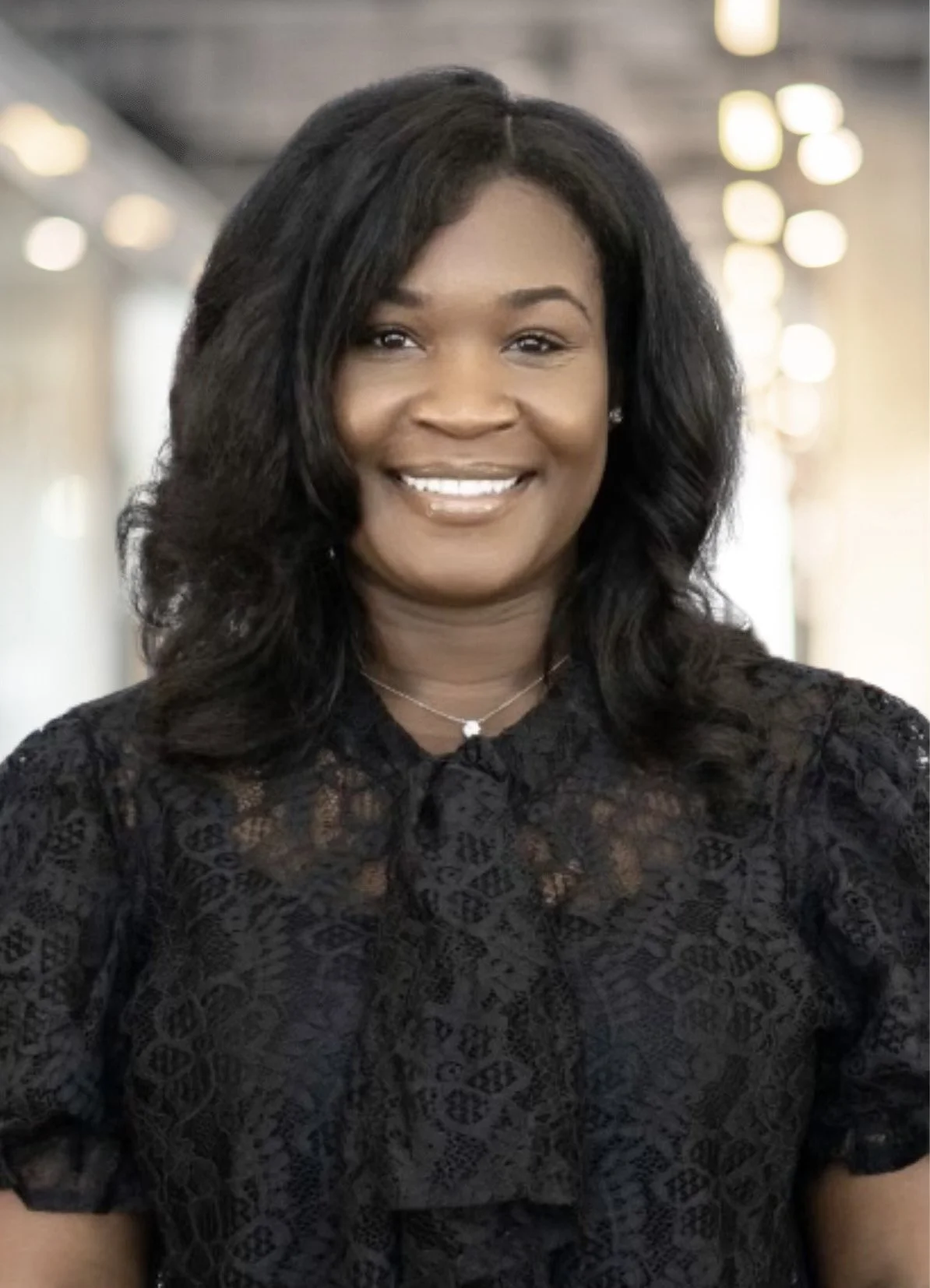 Portrait of a woman with dark wavy hair smiling, wearing a black lace top and a necklace, in a well-lit indoor setting.