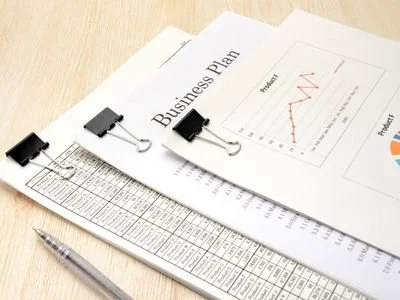 Business documents including a business plan with a graph, a spreadsheet, a ruler, a pen, and binder clips on a wooden desk.