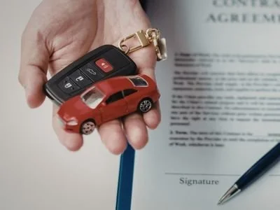 Hand holding a set of car keys with a small red toy car on top, near a document with a signature line and a pen.