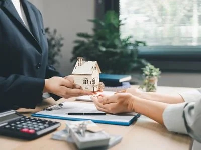 A person handing over a small wooden house model to another person during a real estate transaction at a desk.