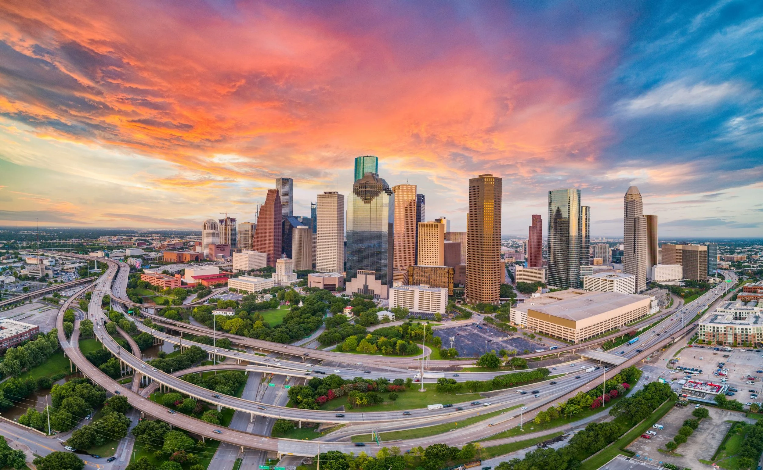 A city skyline with tall skyscrapers during sunset, with pink, orange, and purple clouds in the sky and highway overpasses in the foreground.
