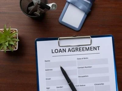 A loan agreement form on a clipboard with a pen, alongside a potted plant, a stapler, and a small blue object on a wooden desk.