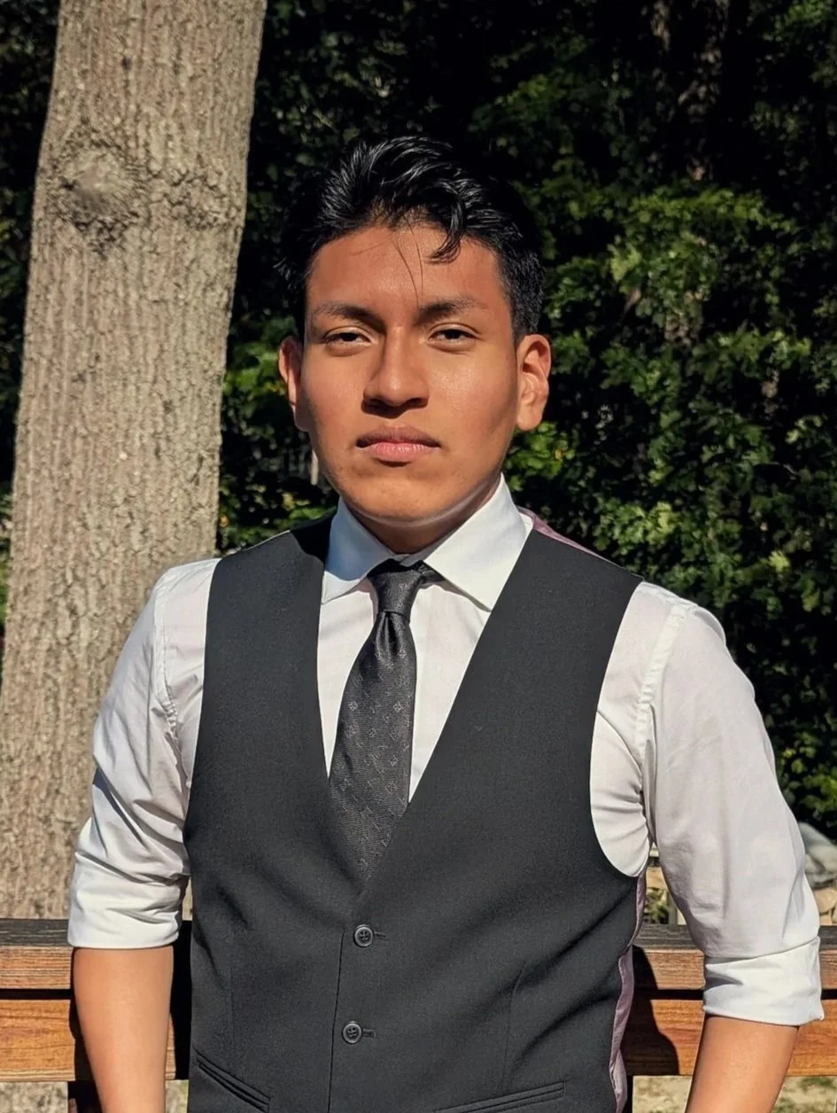 Young man in a white shirt, black vest, and black tie standing outdoors near a tree, with greenery in the background.