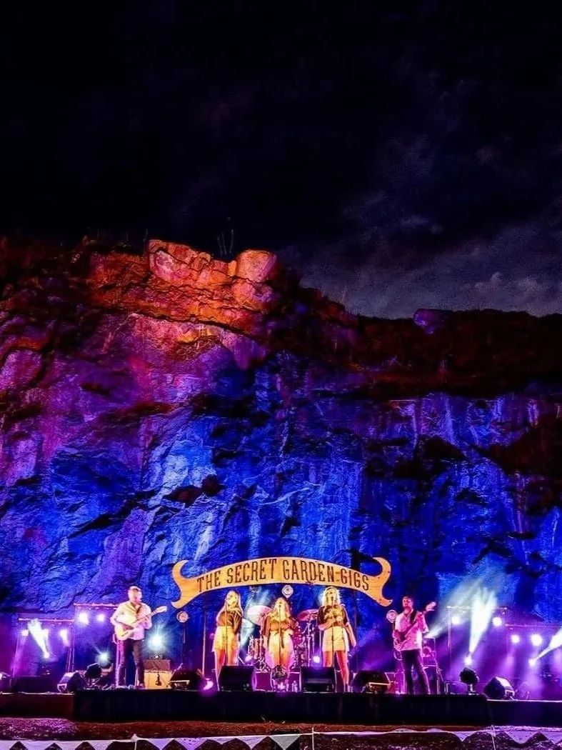 A band performing on stage at night with a rocky cliff background lit with colorful lights, and a banner that reads 'The Secret Garden Gigs'.