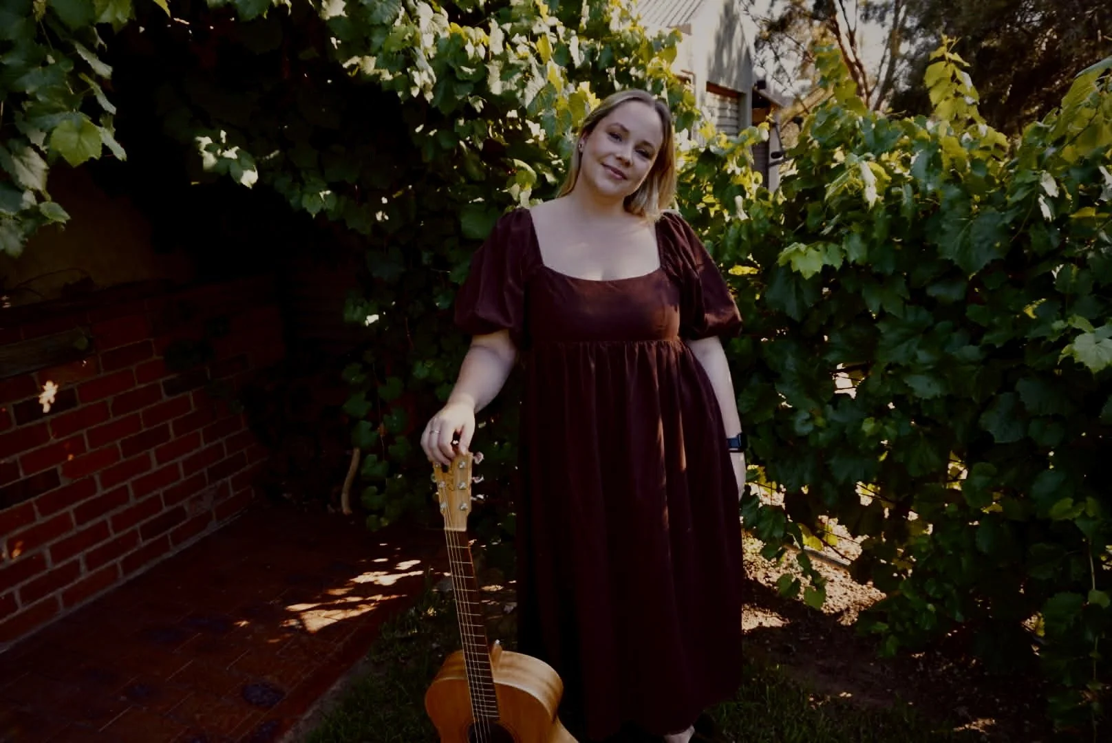 A woman with shoulder-length blonde hair wearing a burgundy dress, standing outdoors near green bushes and a brick wall, holding an acoustic guitar.
