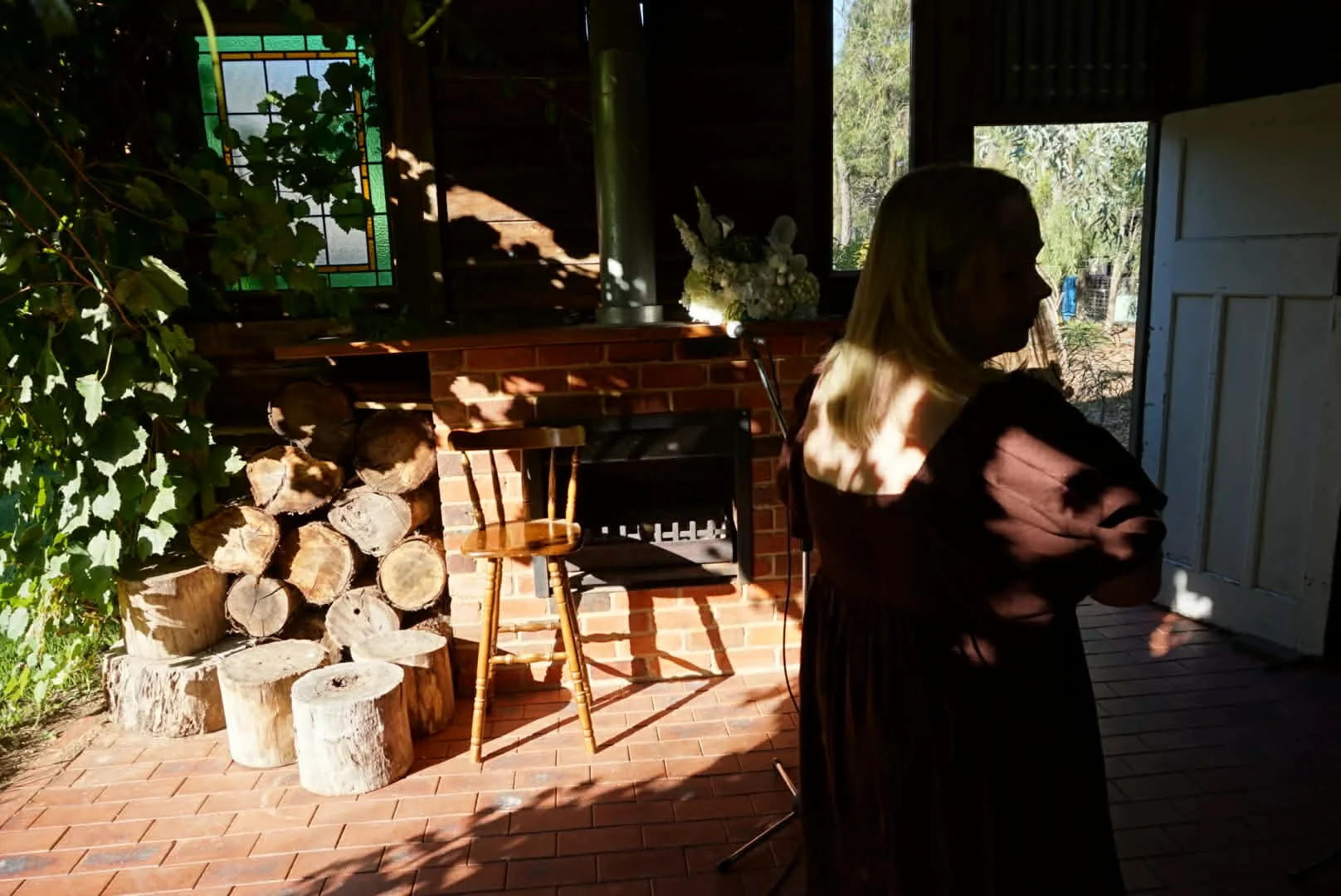 Interior of a rustic house with a brick fireplace, stacked firewood, a wooden chair, a black seat, and a bouquet of white flowers on the mantel. Two women are standing near the open door, one with blonde hair and the other with dark hair. Sunlight filters through the windows, casting shadows.