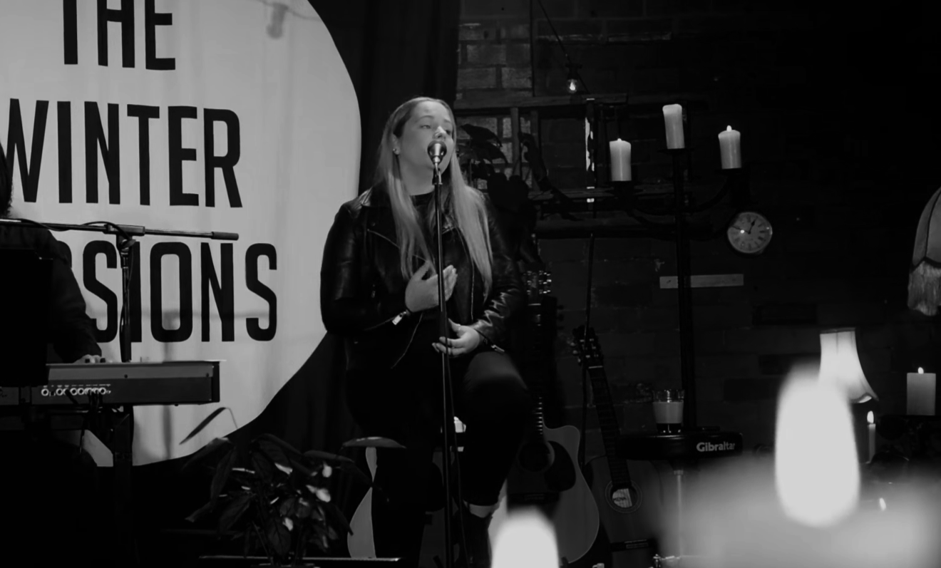 A woman singing on stage with a microphone, seated with her hand over her chest. Behind her is a large banner reading 'The Winter Sessions'. Stage decor includes candles and musical instruments, with a keyboard and guitars visible.