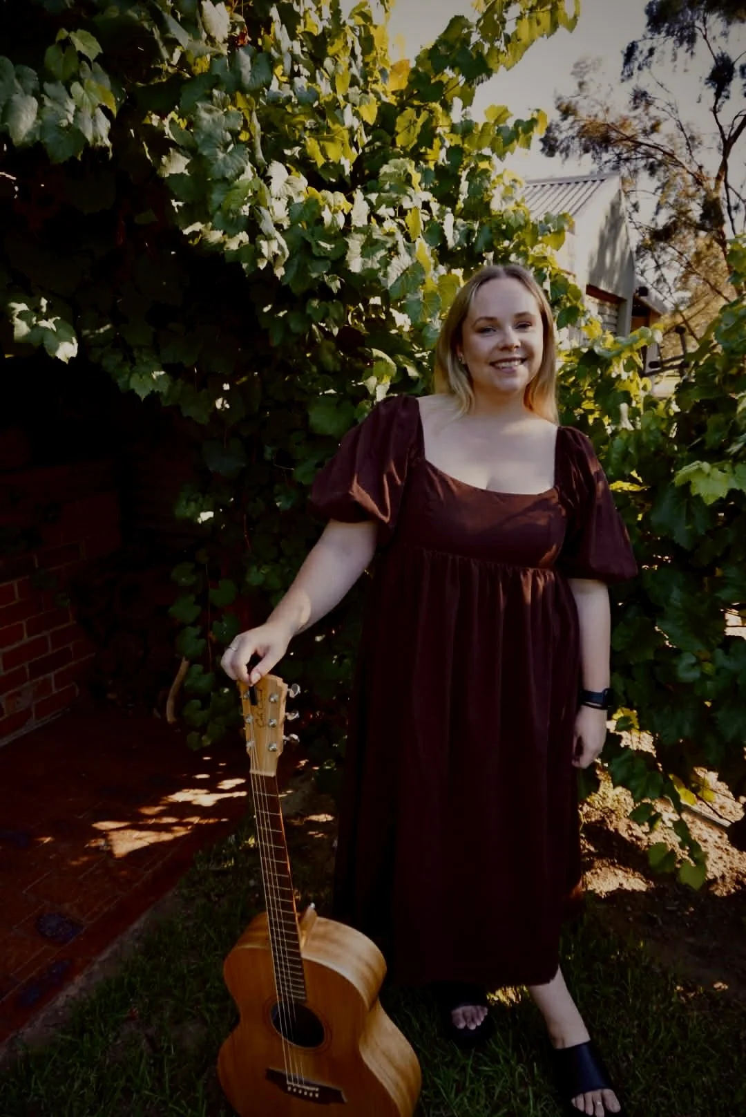 A young woman with blonde hair wearing a dark red dress with puffed sleeves, standing outdoors in front of green leafy bushes, holding an acoustic guitar in her right hand, smiling at the camera.