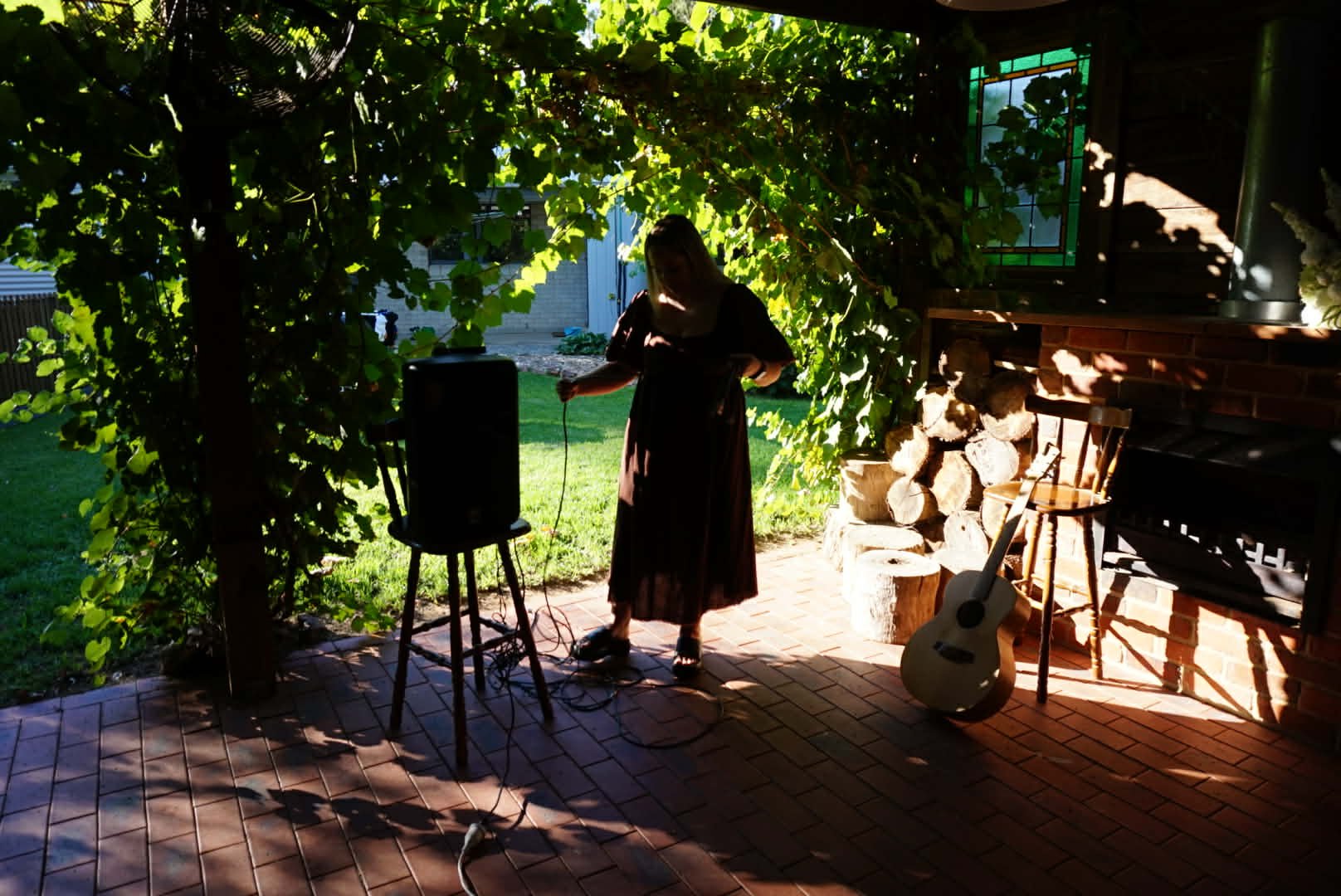 A woman standing on a brick patio outside, under green leafy vines, with a guitar resting against a stack of firewood and a chair nearby. She wears a long dark dress and is adjusting something with a speaker and a table in front of her.
