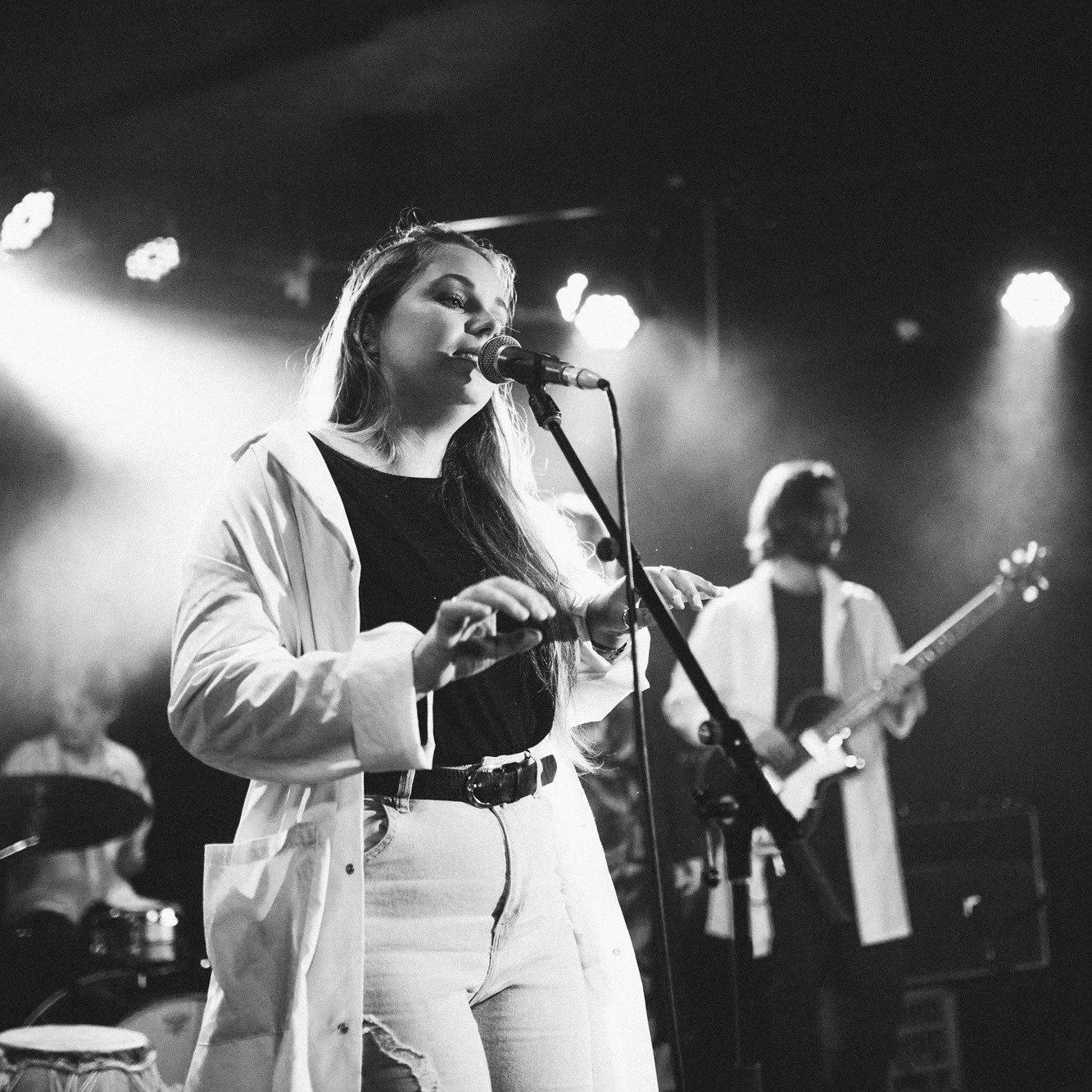 A female musician singing into a microphone on stage, with a guitarist in the background, grayscale concert scene.