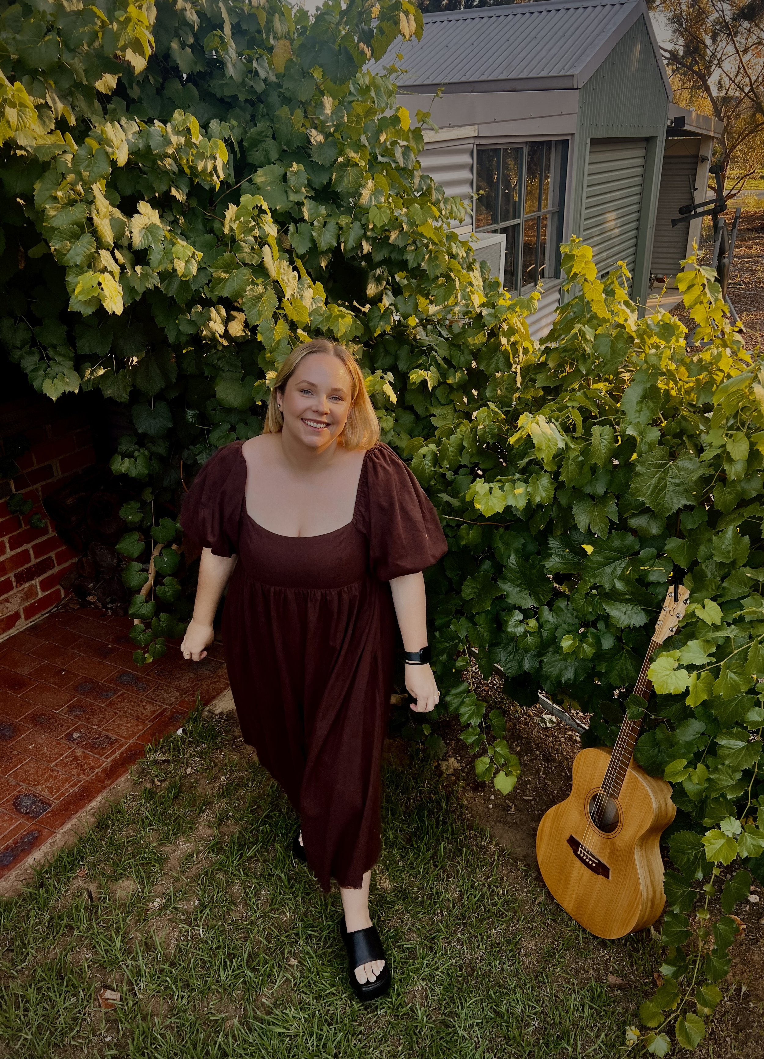 A woman with blonde hair smiling, wearing a maroon dress and black shoes, standing outdoors next to a guitar, surrounded by green foliage and a house with a window and a shed in the background at sunset.