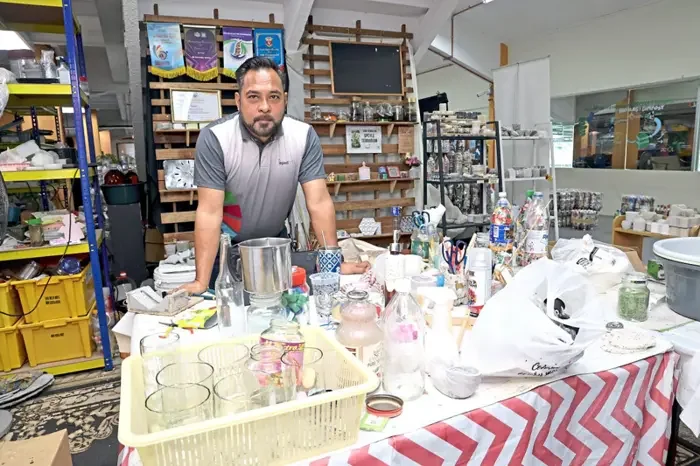 A man stands behind a cluttered table filled with various bottles, glasses, and household items in a shop or craft store, with shelves and displays in the background.