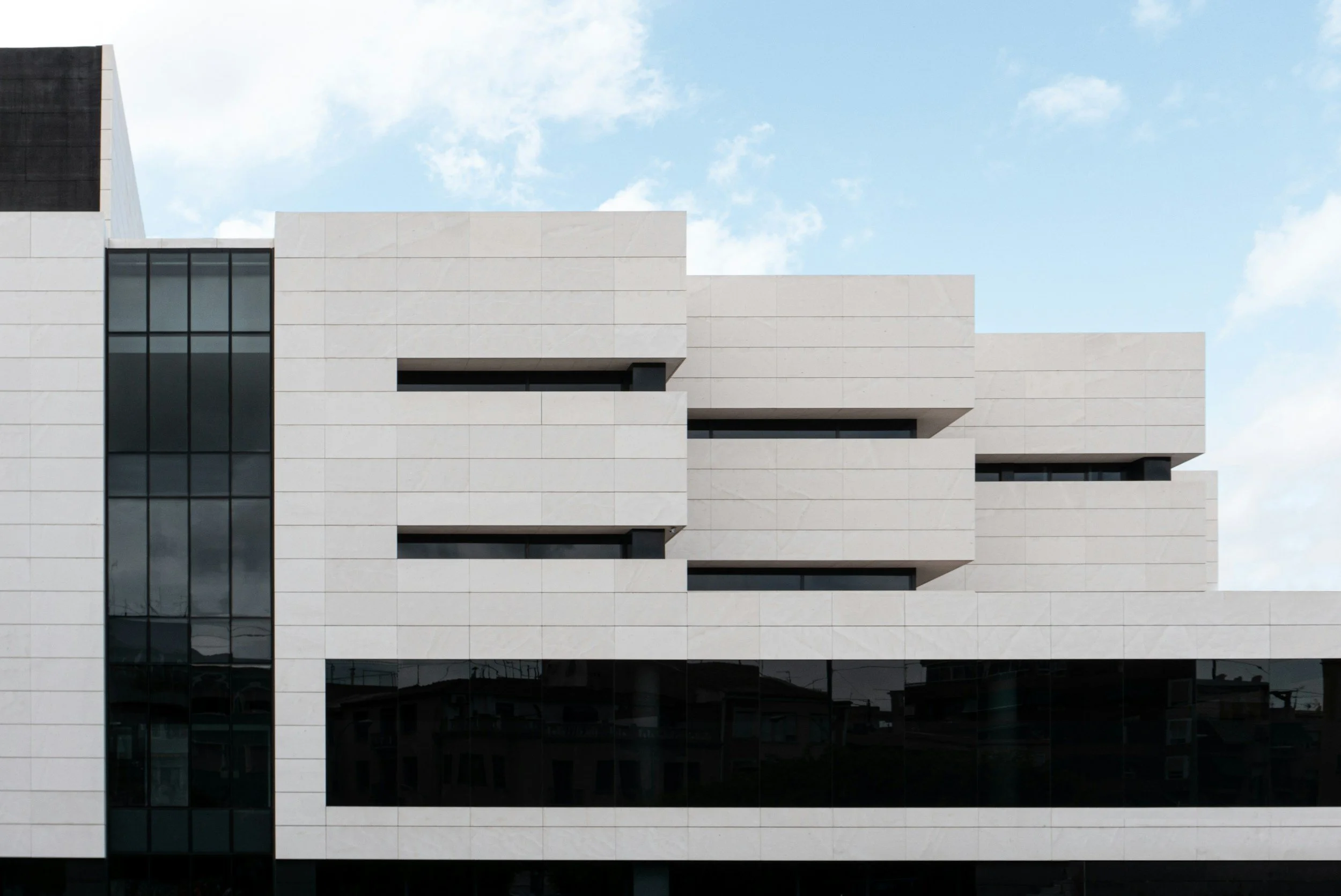 Modern multi-story building with white facade and black window accents, sky with scattered clouds in the background.