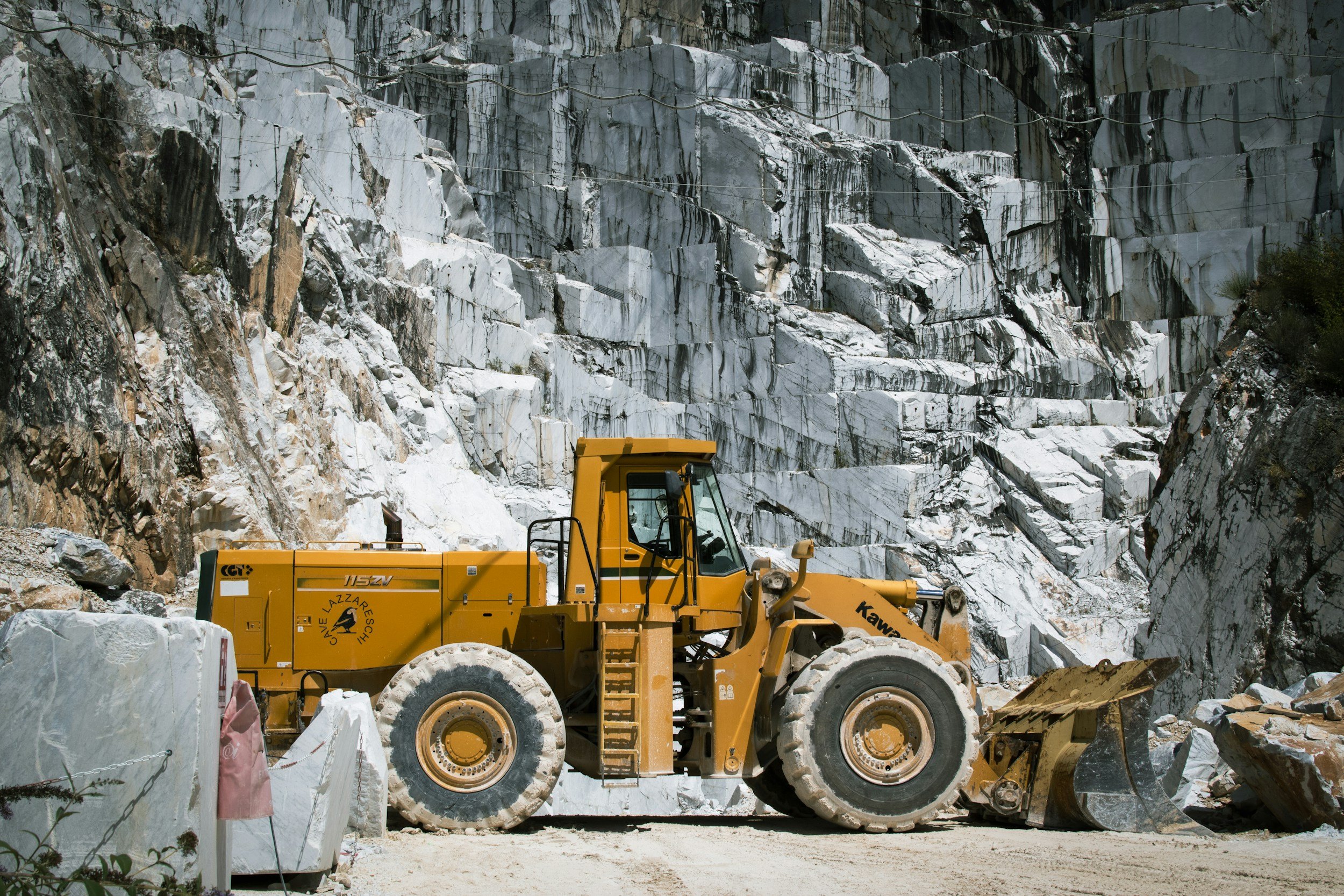 A yellow construction vehicle working at a marble quarry, with large white marble rocks and a rocky cliff face in the background.