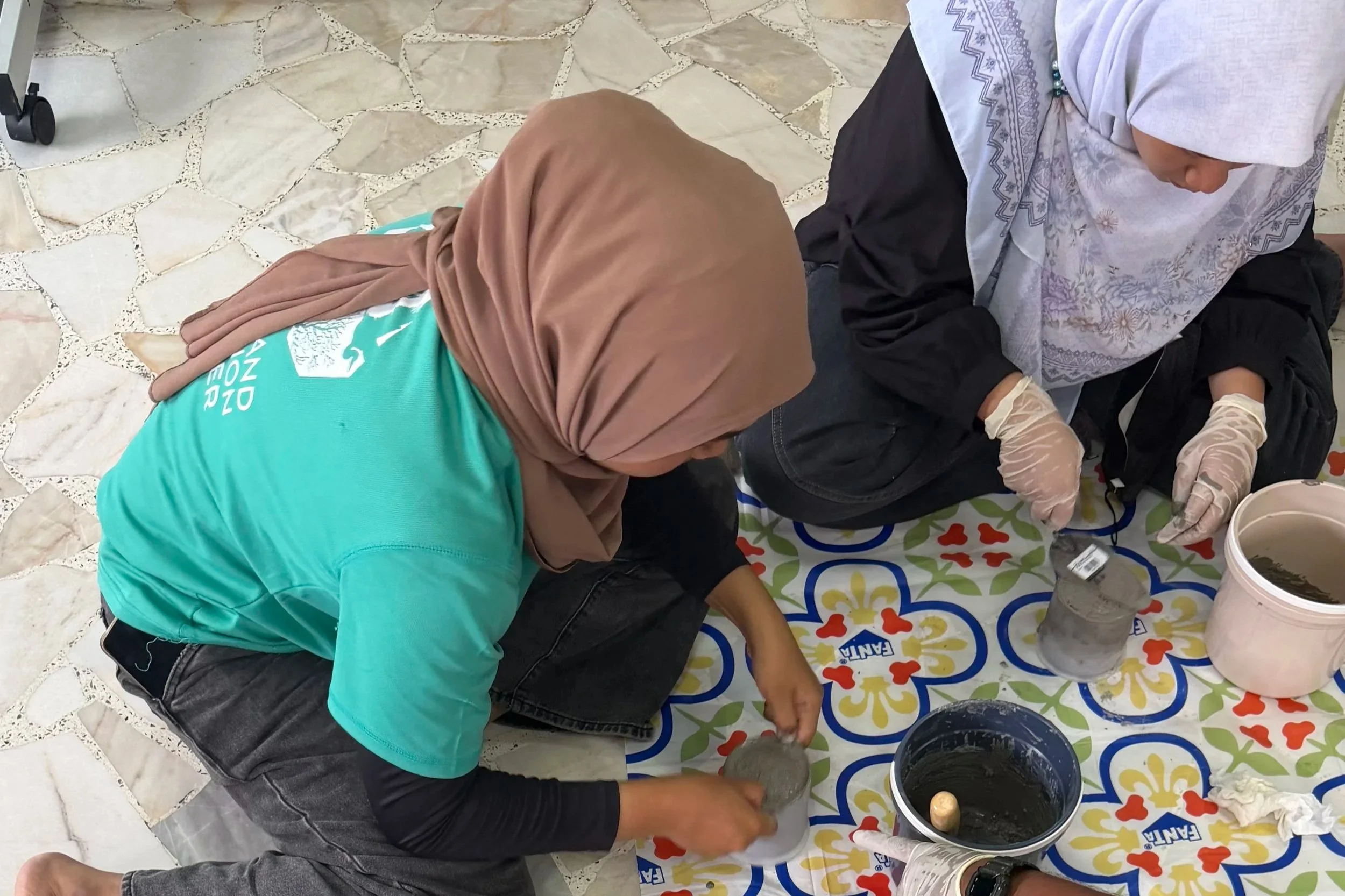 Two women wearing headscarves and gloves are kneeling on a colorful patterned cloth on the floor, working with wet cement, surrounded by buckets of cement and tools.