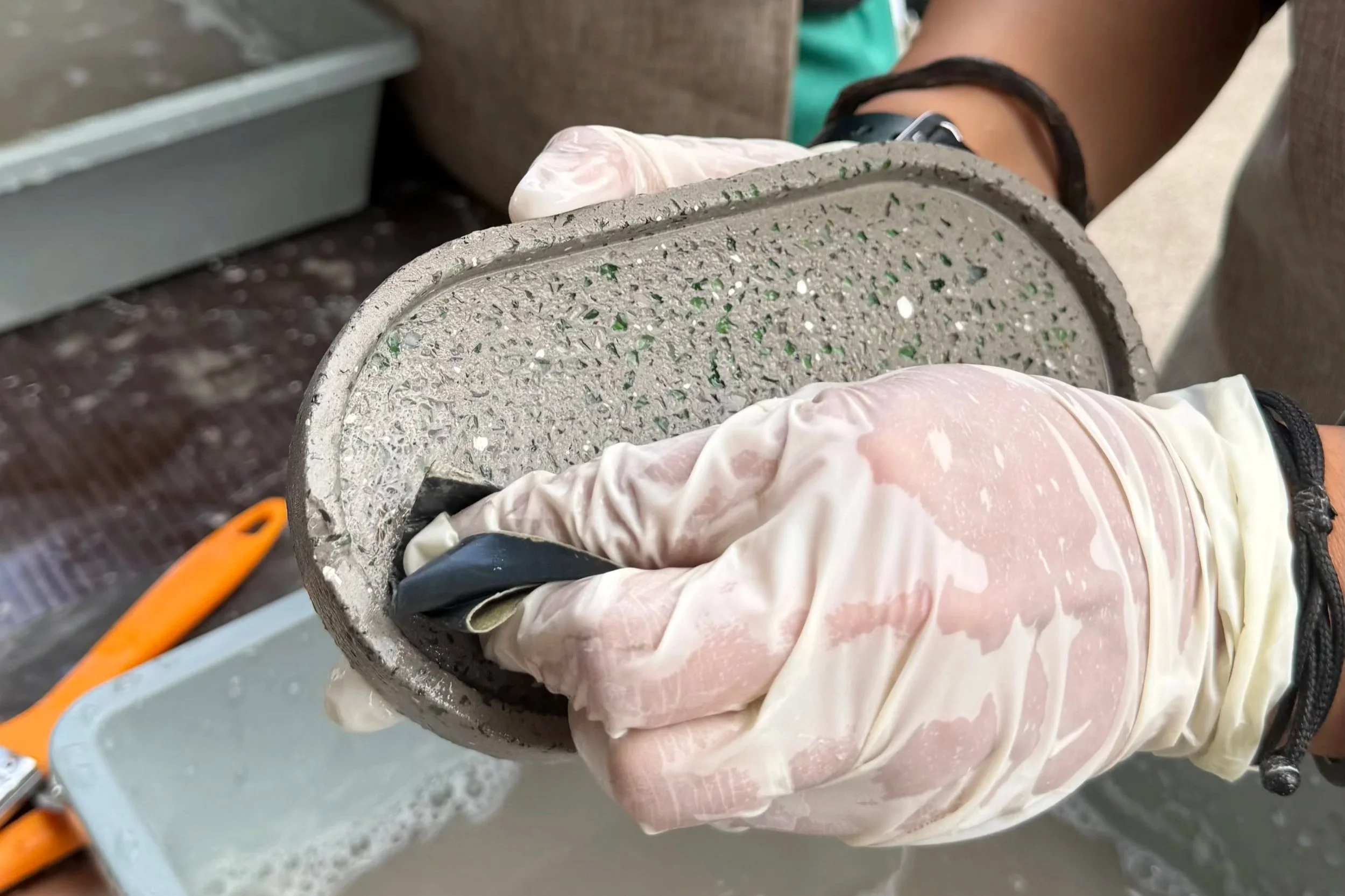 Person wearing gloves holding a concrete sample with small stones and green inclusions, in a laboratory or industrial setting.