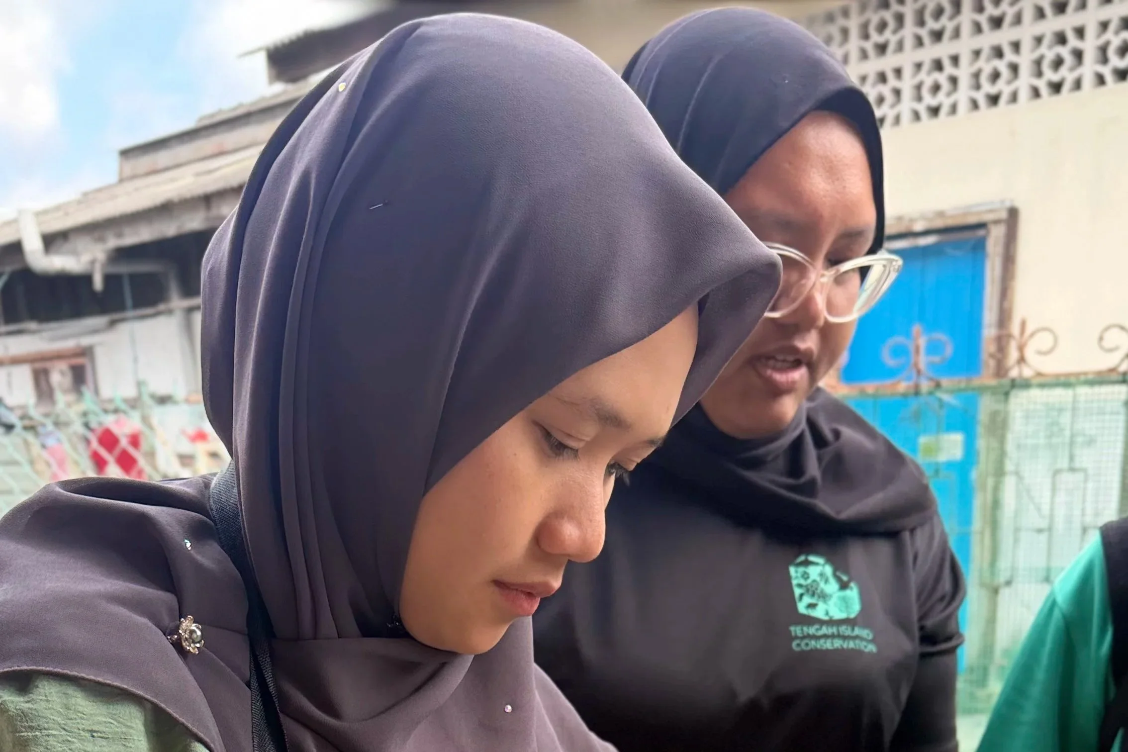 Three women wearing hijabs and dark clothing, engaging in an outdoor activity, with blurred buildings and fence in the background.