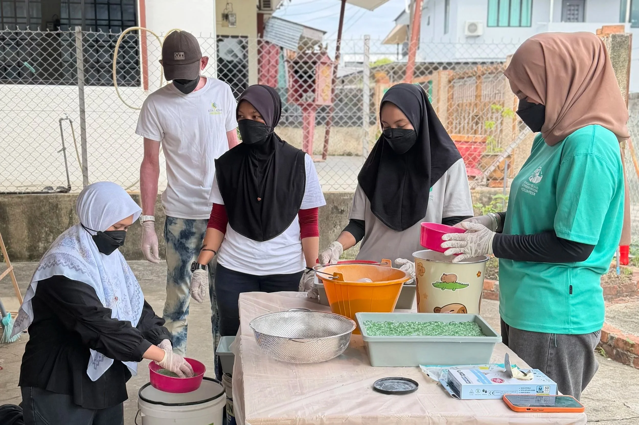 People wearing face masks and gloves standing around a table with bowls and supplies outside, possibly participating in a community activity or volunteering.