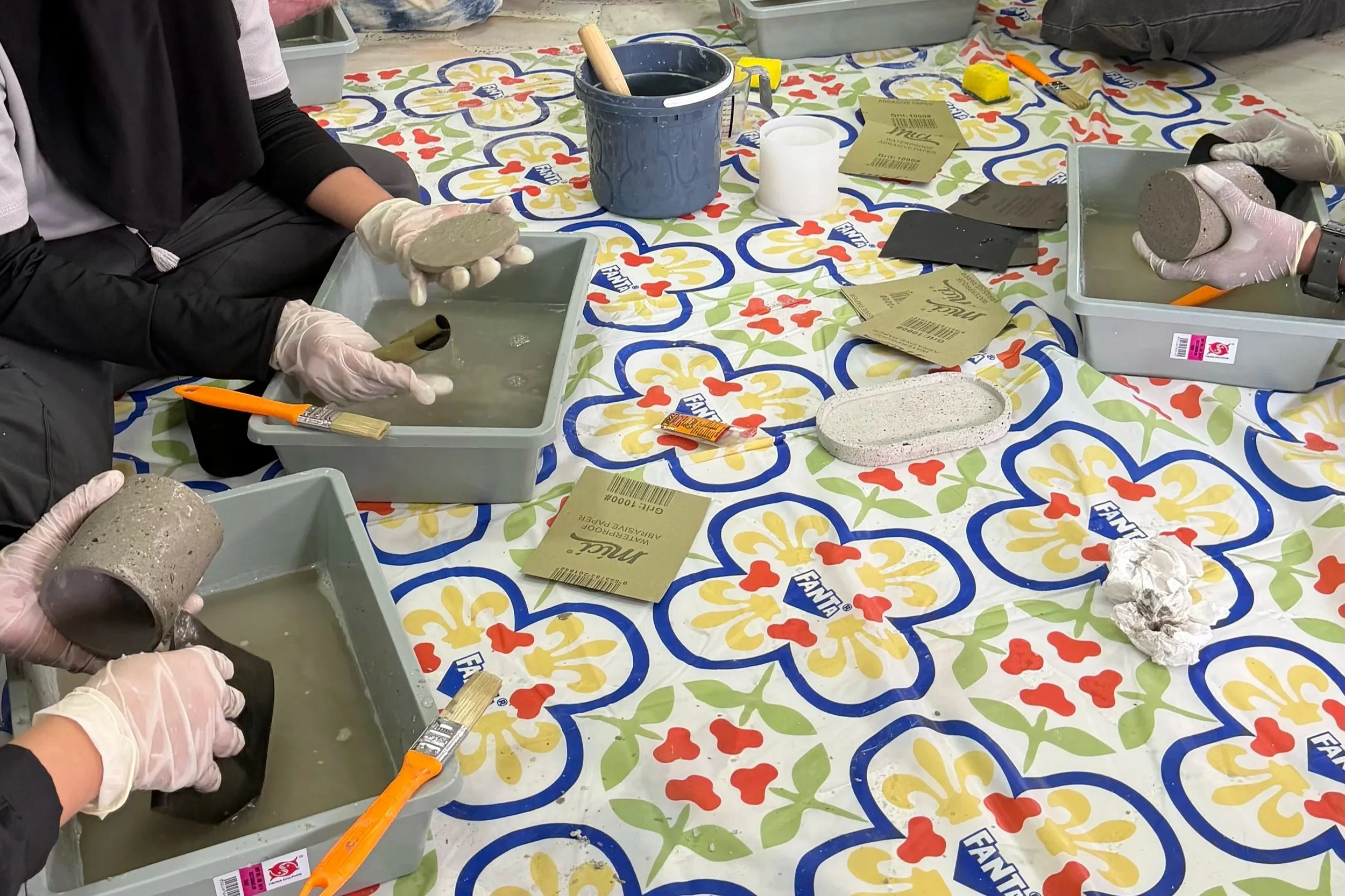Individuals wearing gloves working on ceramic tile crafts at a colorful table, with various tools, brushes, and materials like tiles and molds present.