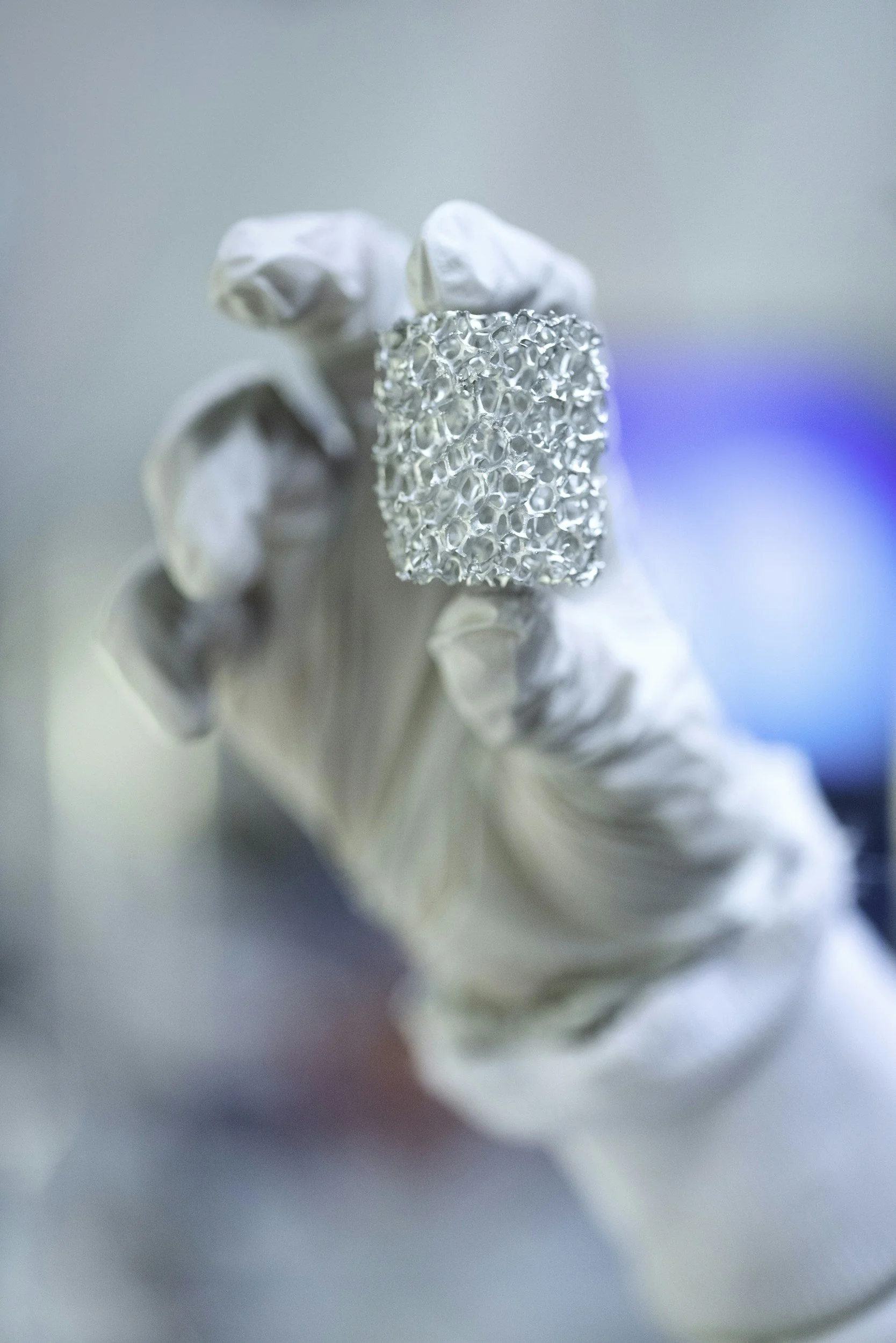 Close-up of a metallic, textured object held by a hand wearing a white glove, with a blurred background featuring shades of blue, green, and red.