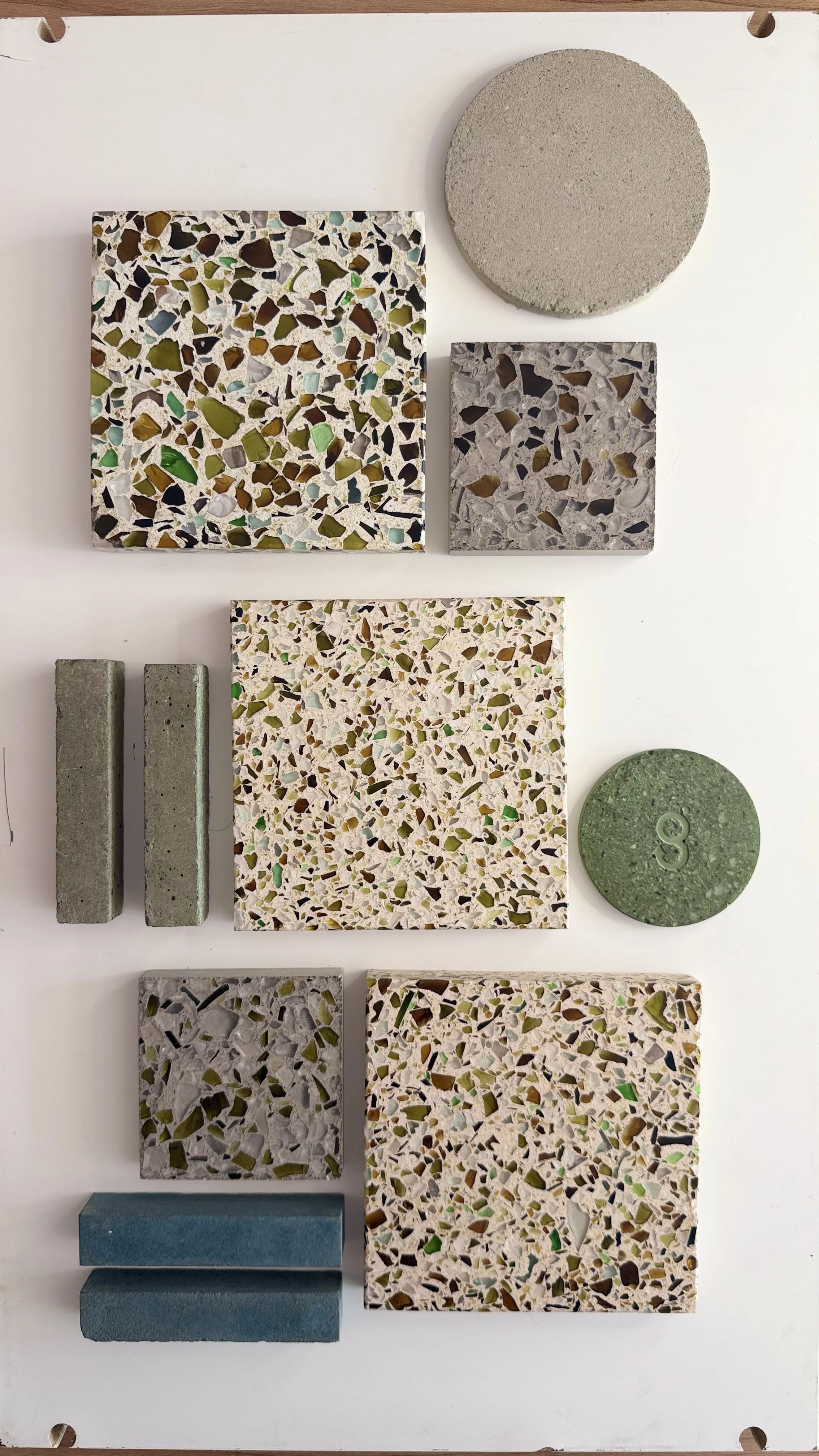 Collection of various tiles and materials, including square terrazzo tiles with multicolored chips, two rectangular concrete tiles, a gray textured circular tile, a light green circular tile, and blue rectangular tiles, on a white surface.