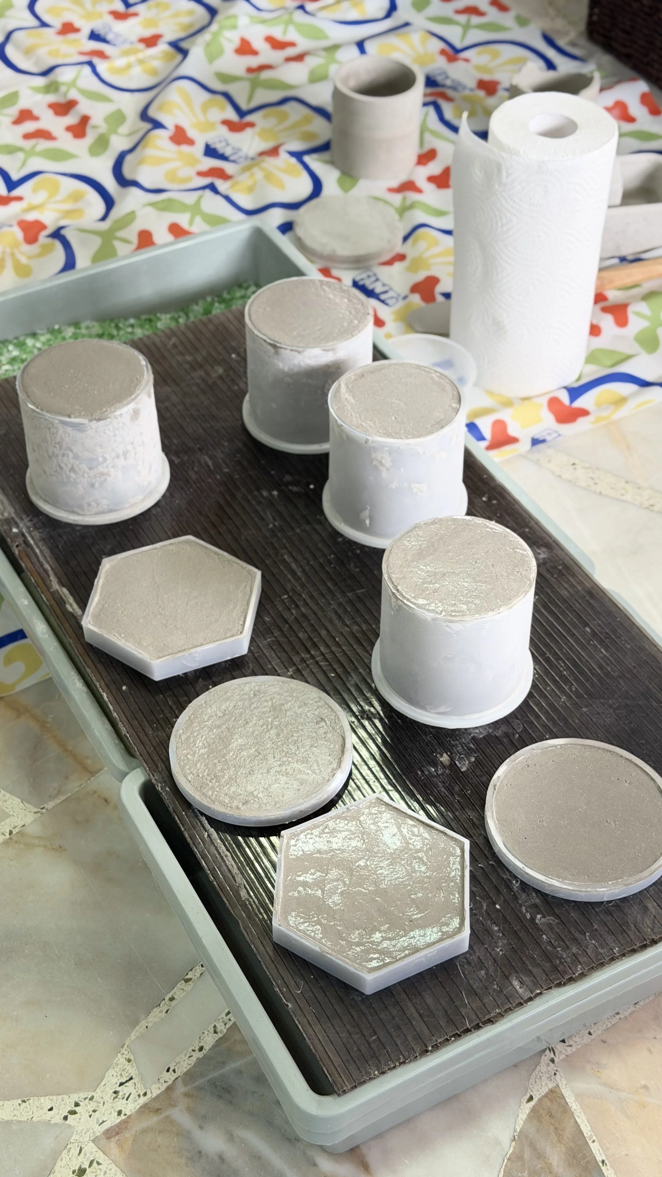 Assorted molds and supplies for making handmade soap, including cylindrical and hexagonal molds, on a plastic-covered table with a colorful cloth in the background.