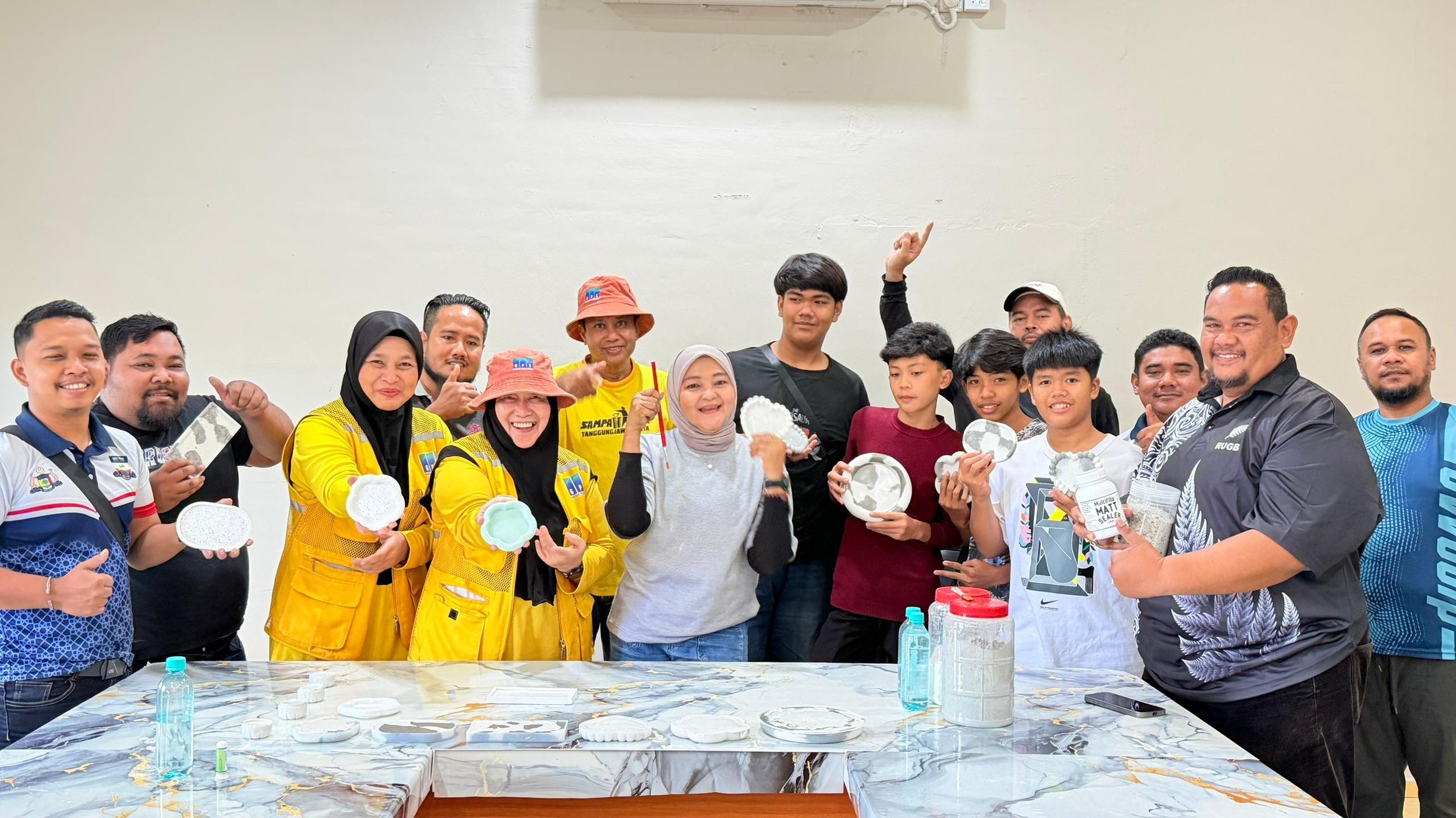 Group of people smiling and holding various ceramic and clay items in an indoor setting, with a marble table in front and a plain wall background.