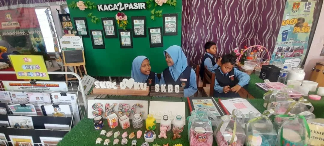 Girls in school uniforms sitting at a table with crafts and decorative items at a market or school fair, with a green backdrop and various posters and displays.