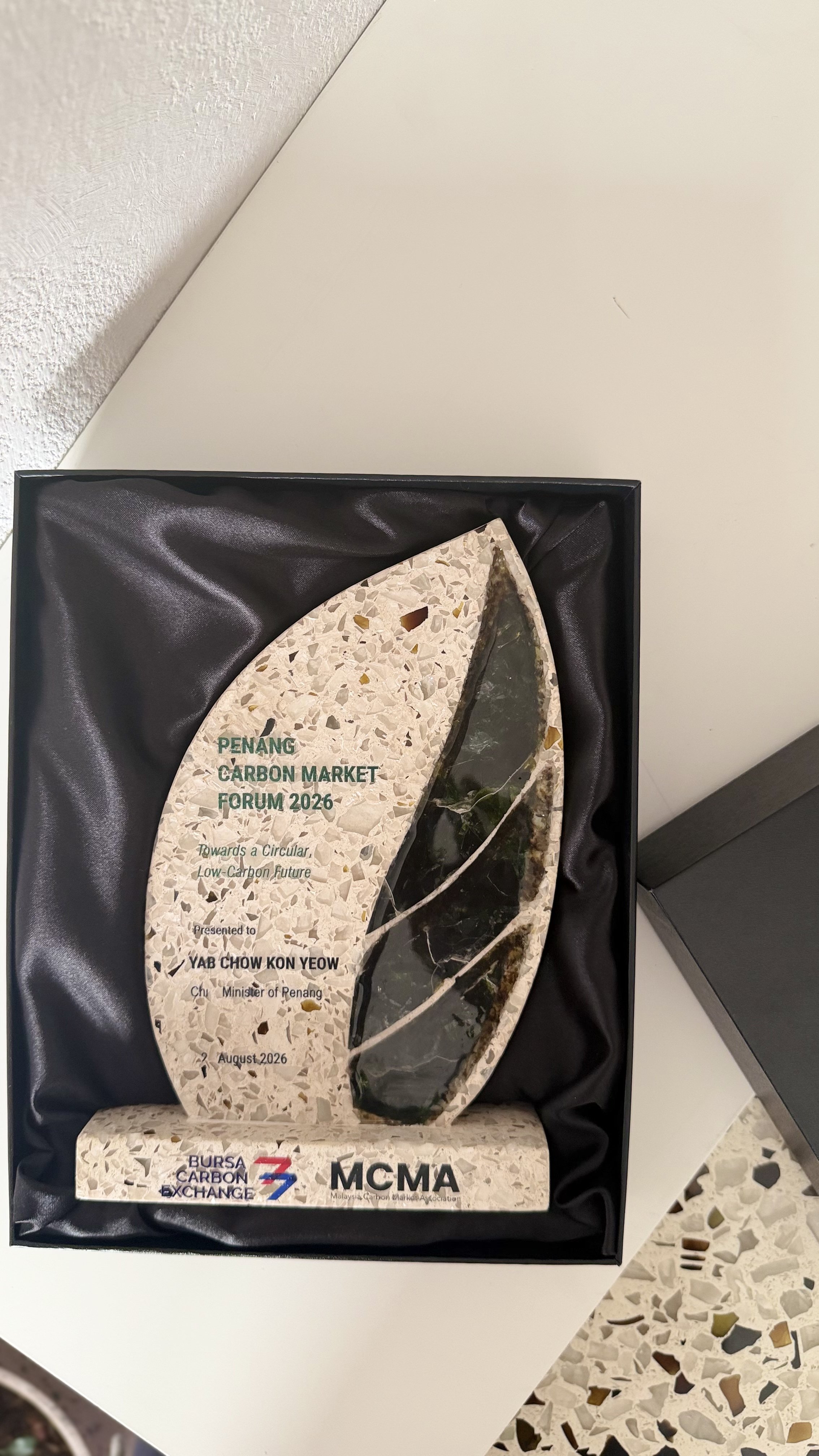 A decorative award plaque with an abstract design, presented at the Penang Carbon Market Forum 2026, placed inside a black box with a satin lining, on a white table with a textured wall in the background.