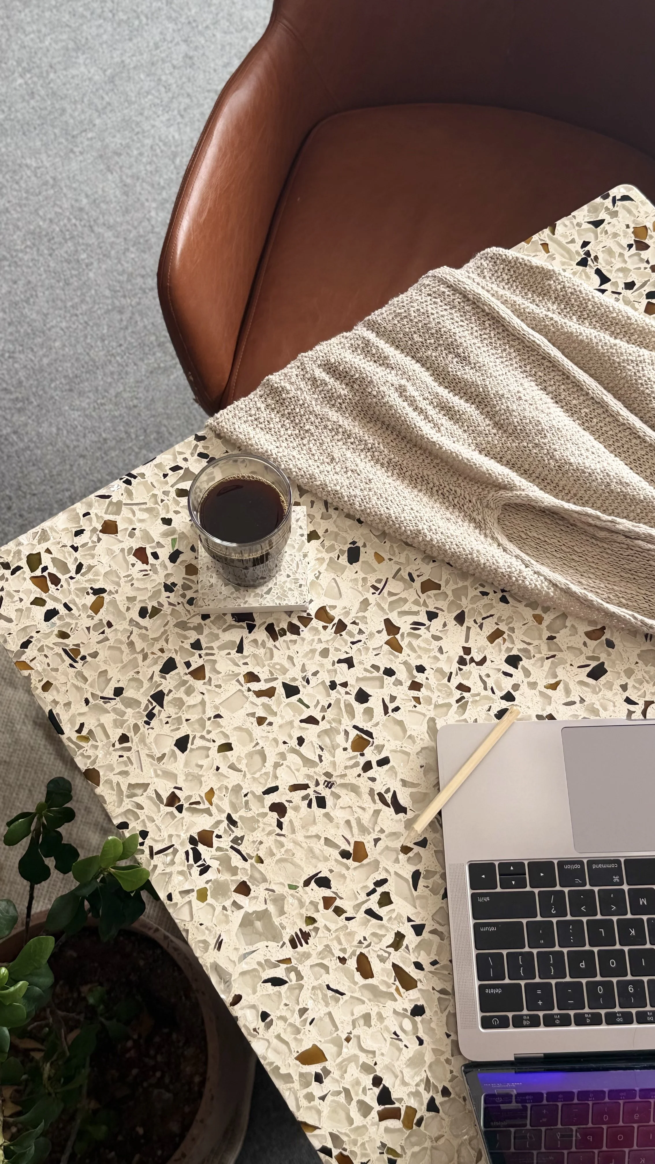A terrazzo table with a glass of dark soda, a silver laptop, a beige textured cloth, a pencil, and a small green potted plant.