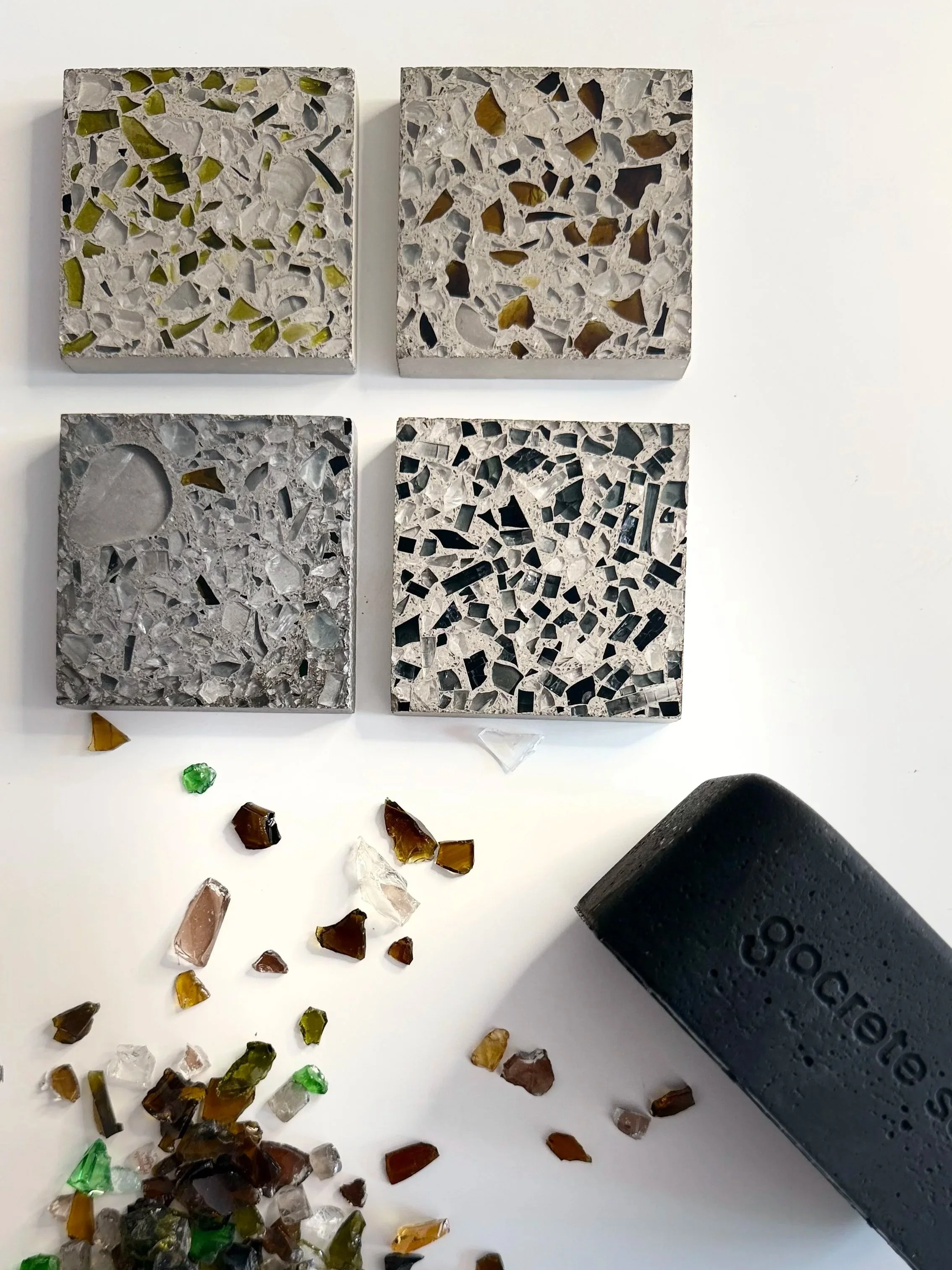 Four mosaic samples with different types of stone and glass chips, arranged in two rows on a white background. Scattered loose mosaic chips and a black portable speaker are in the foreground.