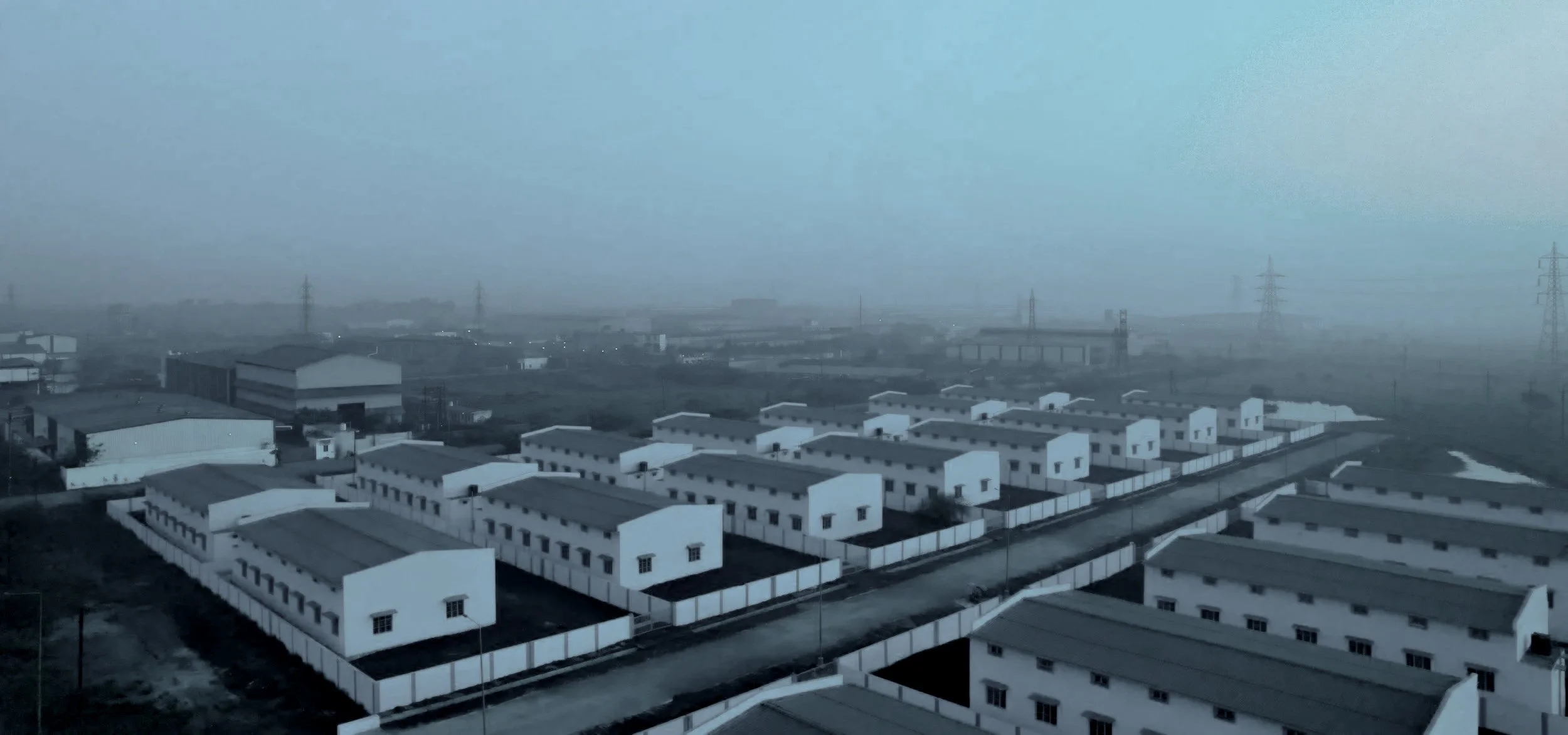 A foggy landscape showing a rural area with multiple white, two-story residential buildings and industrial warehouses. Power lines and pylons are visible in the background.