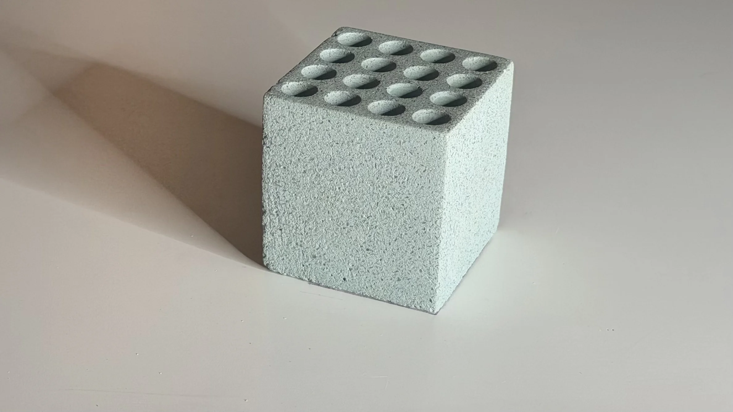 A light gray cement brick with ten circular holes on the top surface, casting a shadow on a white background.
