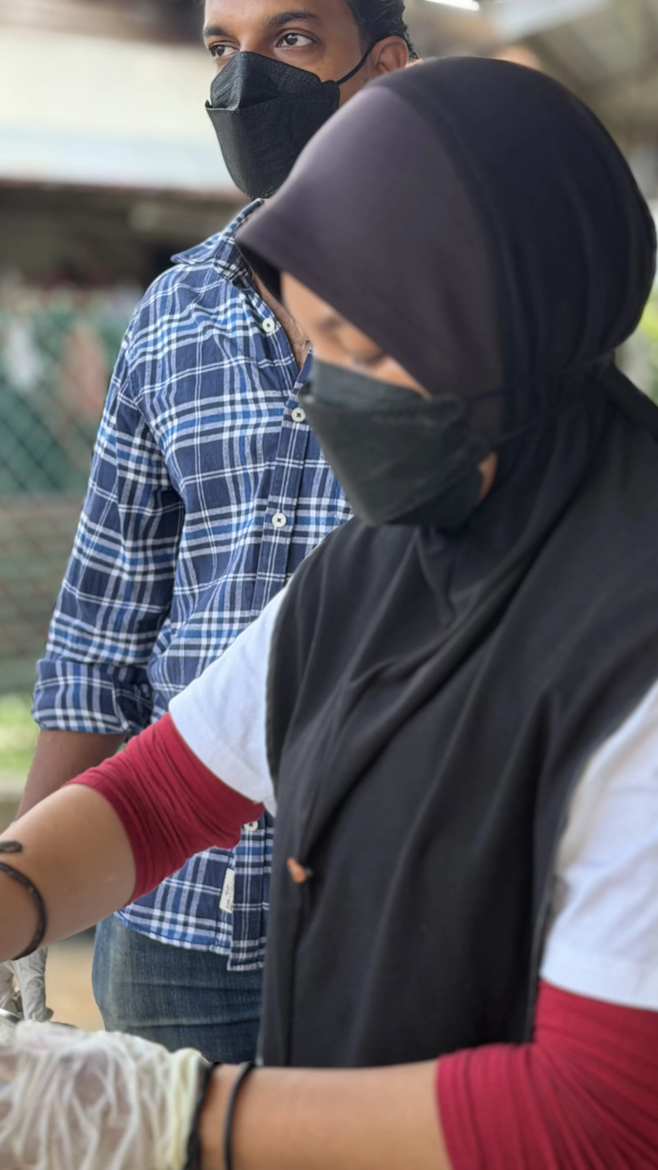 A young man and woman wearing masks, with the man wearing a blue checked shirt and the woman a black hoodie with red and white sleeves, possibly working together on a project outdoors.