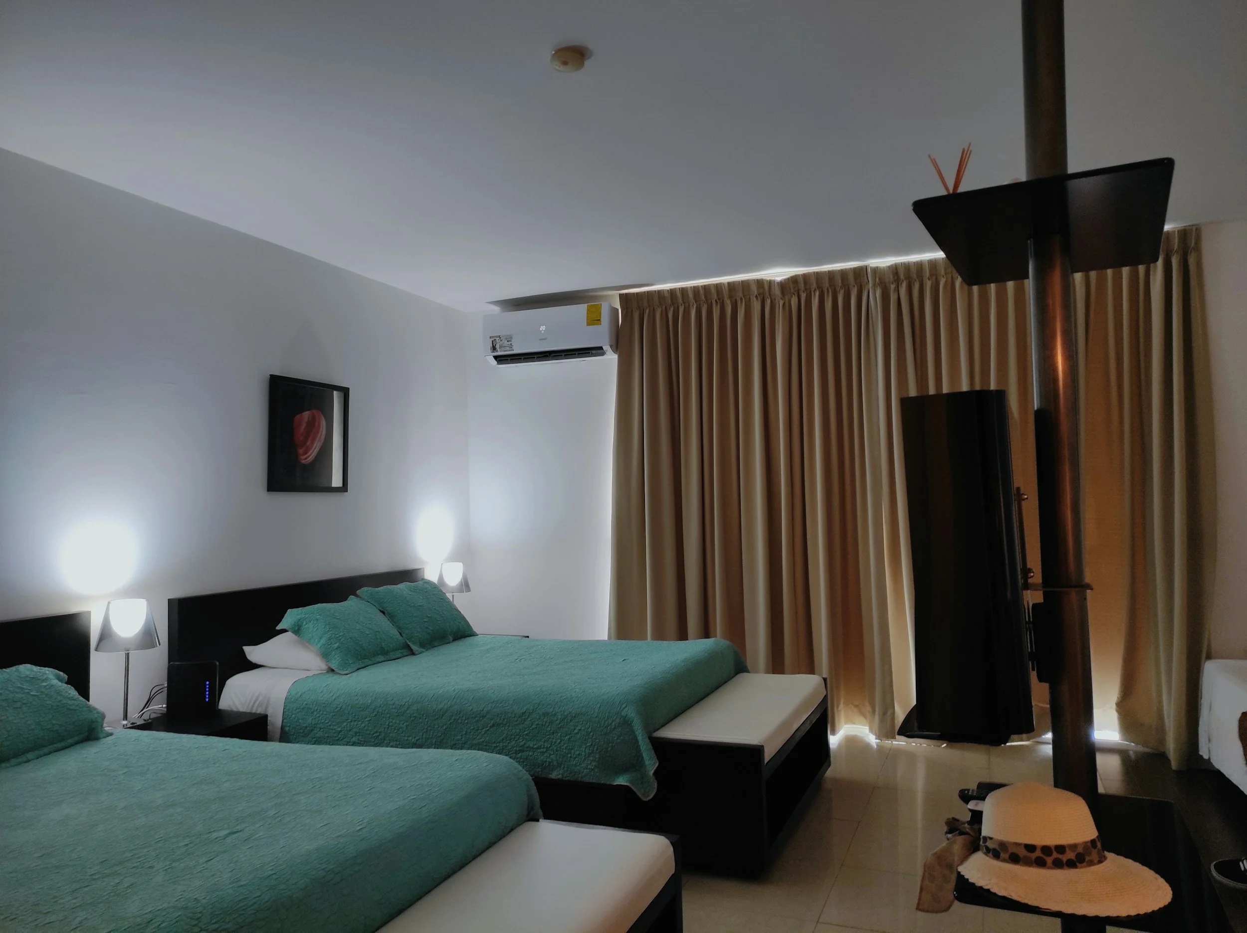 Hotel room with two beds, each with green bedspreads and pillows, beige curtains, a wall-mounted air conditioner, a framed artwork, two bedside lamps, a TV on a stand, and a fedora hat on a small table.