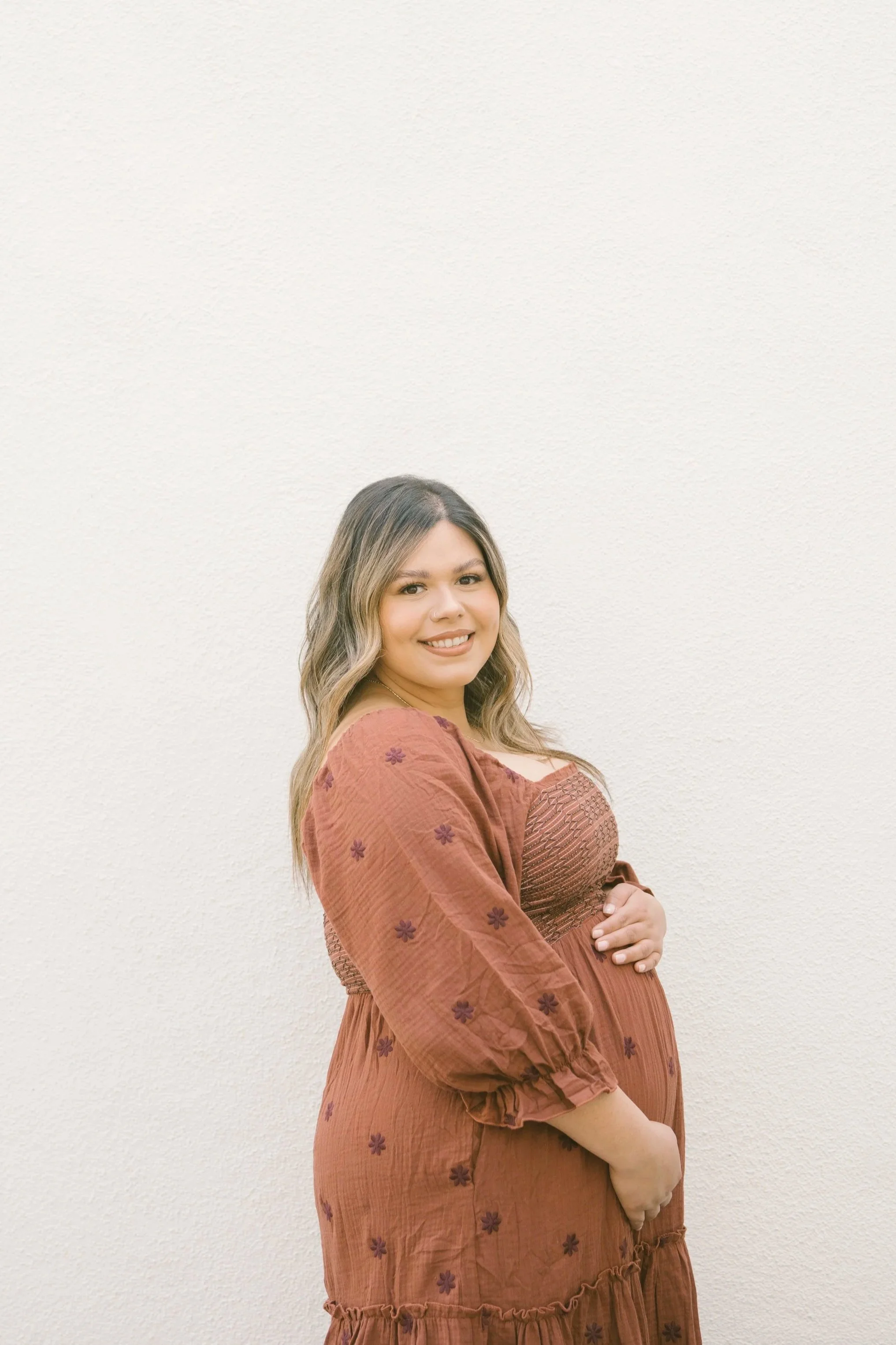 A pregnant woman with wavy light brown hair, wearing a rust-colored dress with embroidered flowers, standing against a plain white wall, smiling and gazing at the camera.