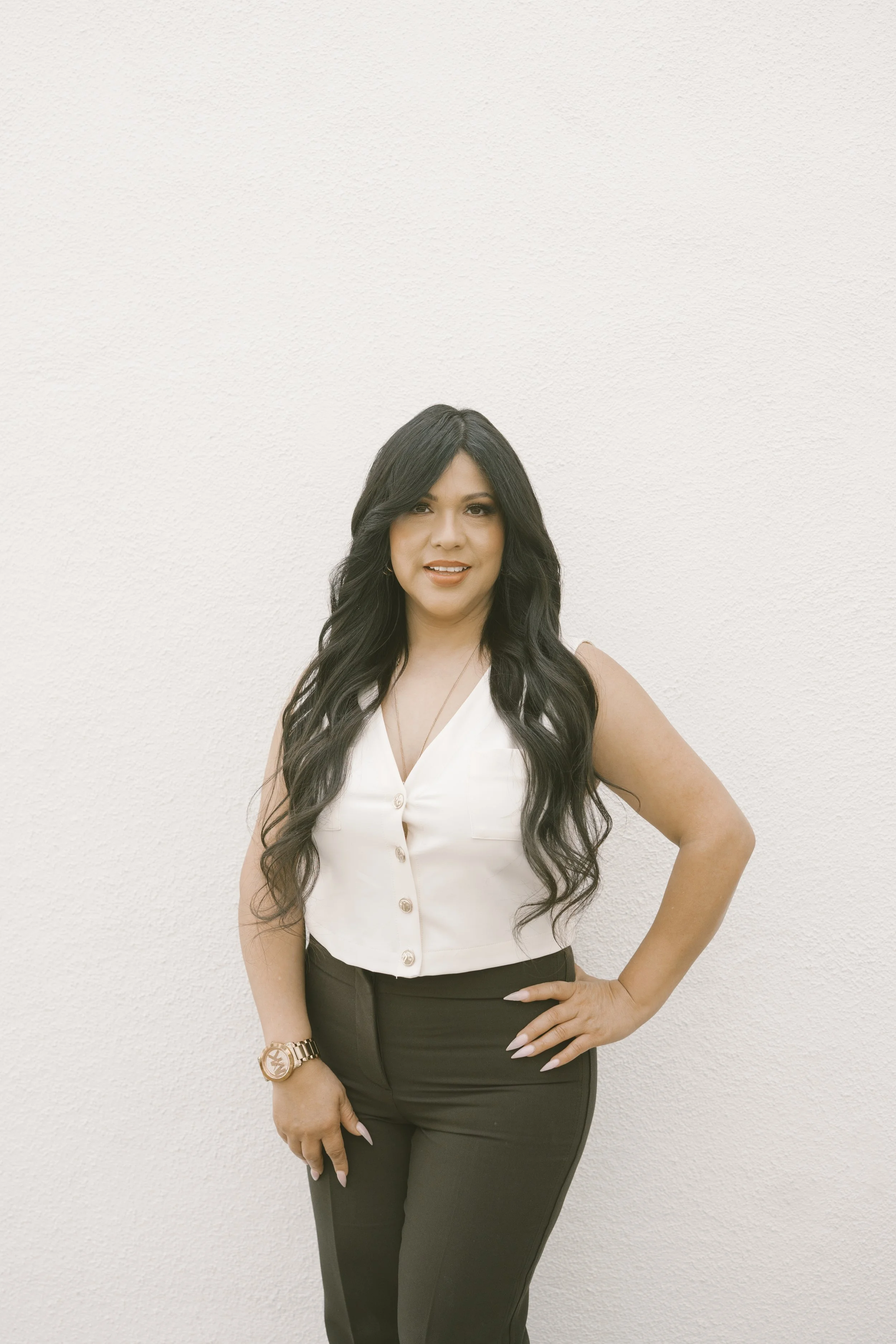 A woman with long black wavy hair, wearing a sleeveless white button-up top and black pants, standing against a plain white wall, with one hand on her hip.